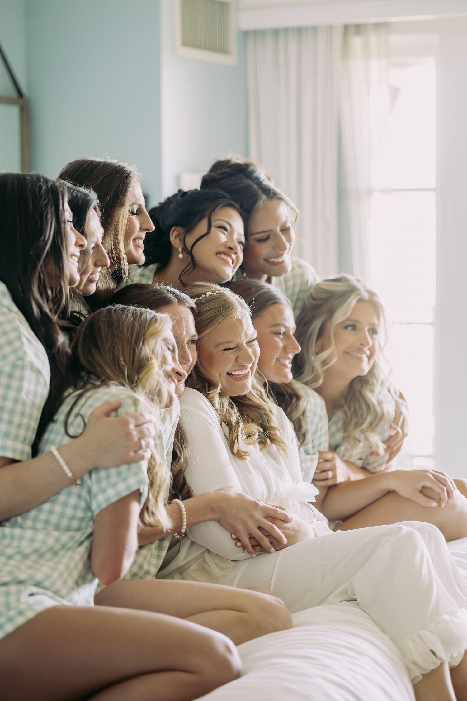 Bride surrounded by bridesmaids and flower girls in matching green gingham pajamas laughing together.