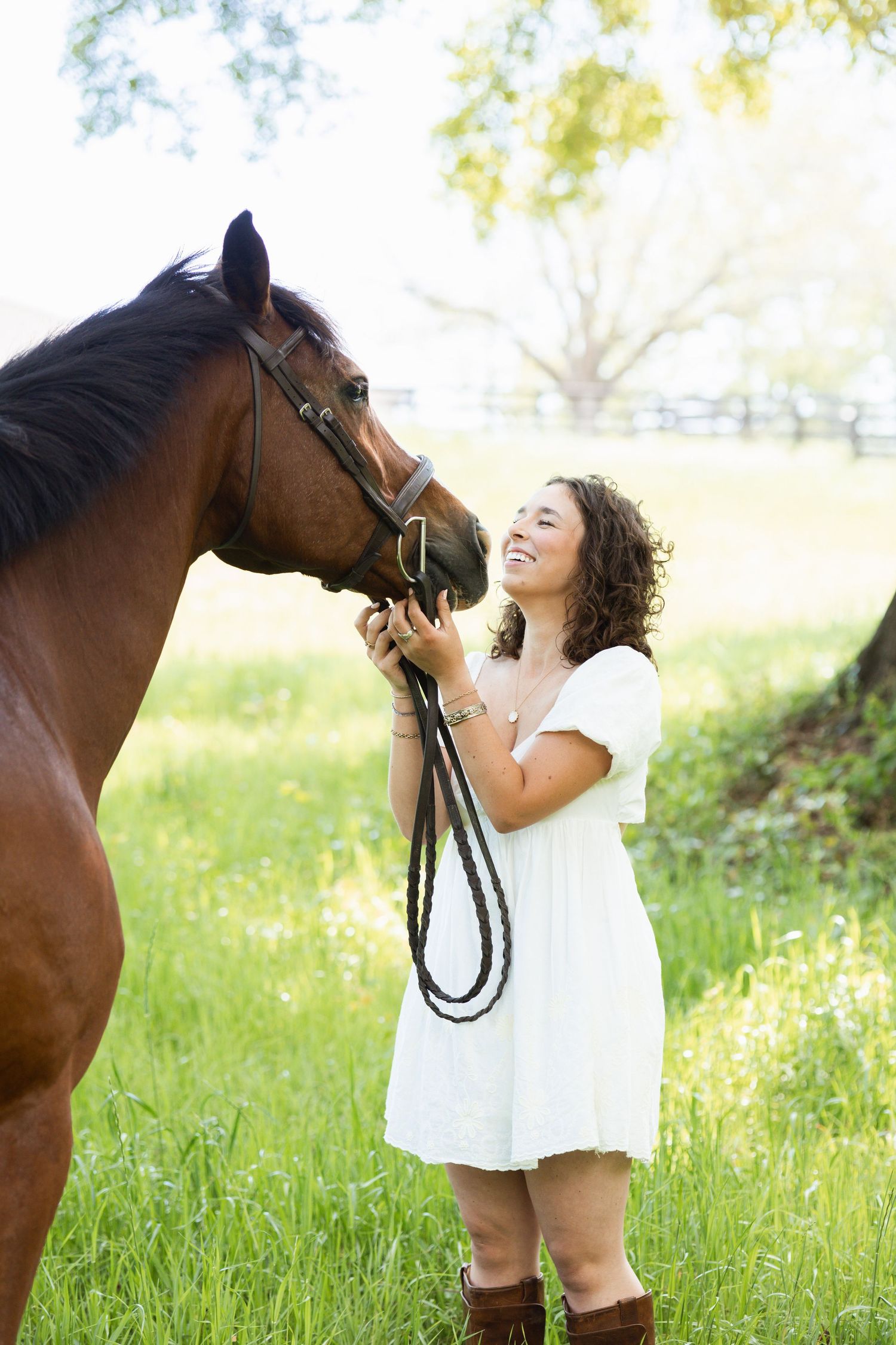 Happy young woman in white dress holding brown horse by bridle in sunny green meadow.