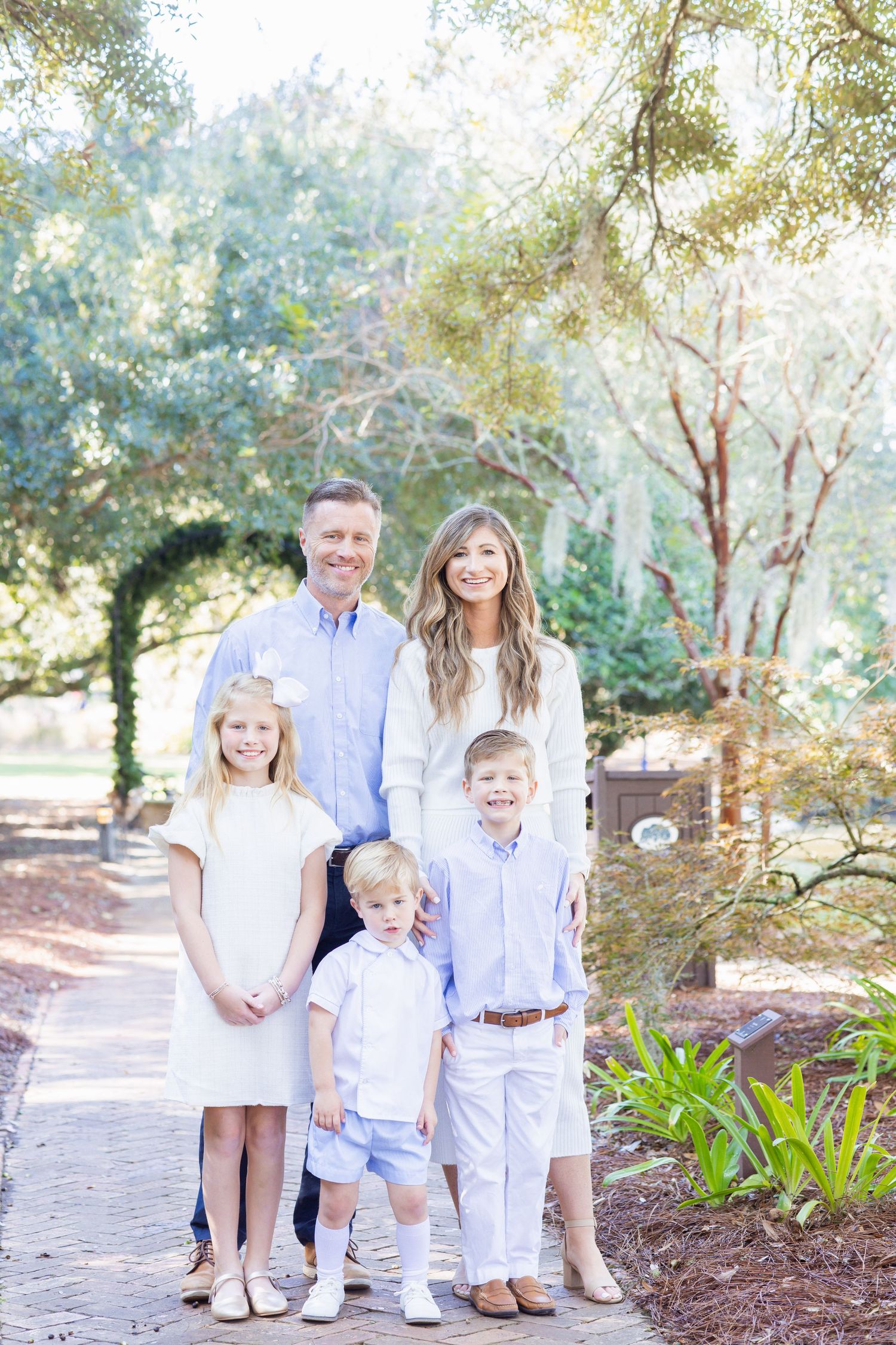 A happy family of five poses on a brick path surrounded by lush green trees, dressed in coordinating light outfits.