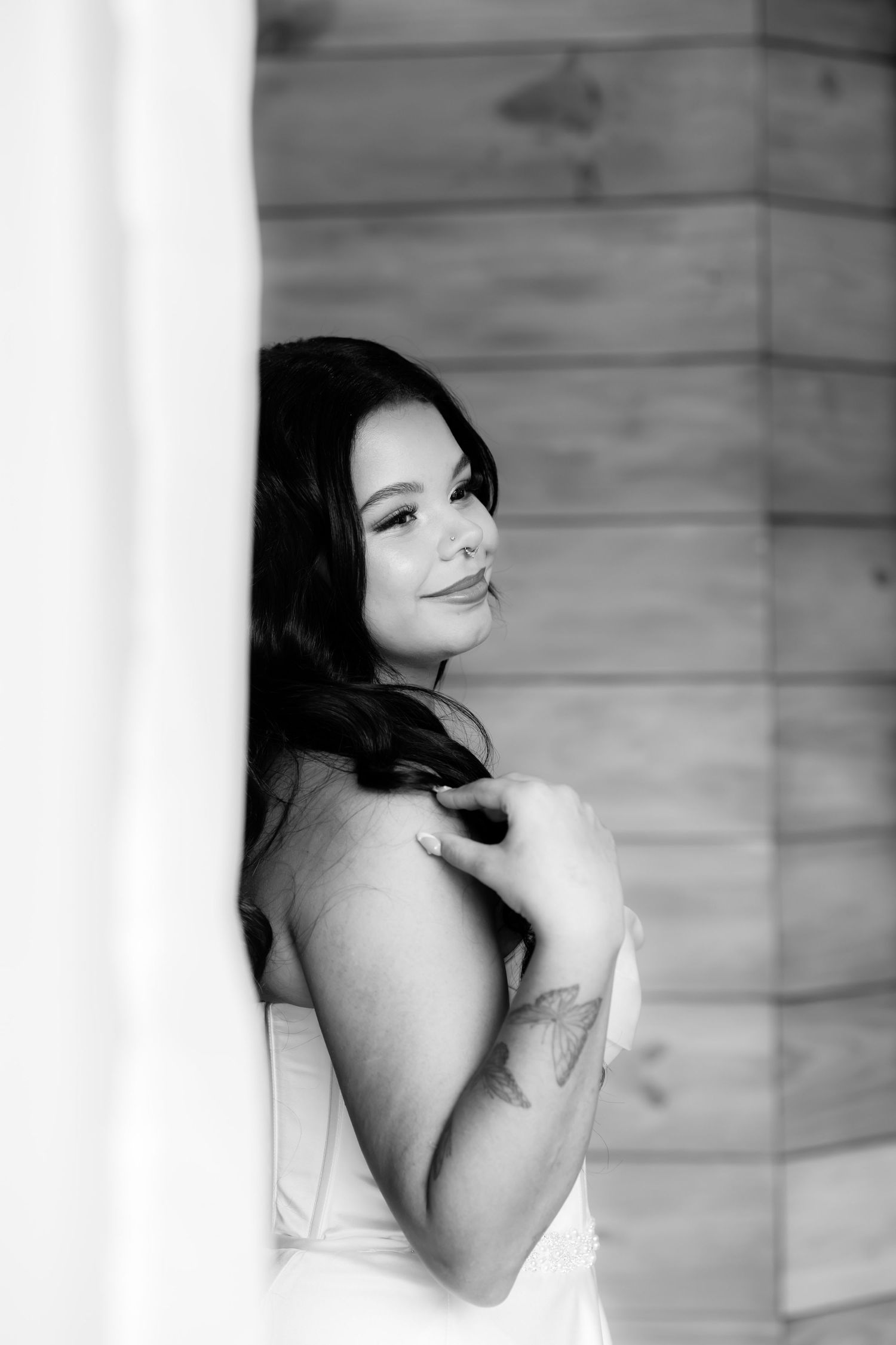 Black and white portrait of dark-haired bride in strapless gown with tattoo, leaning against wood wall.