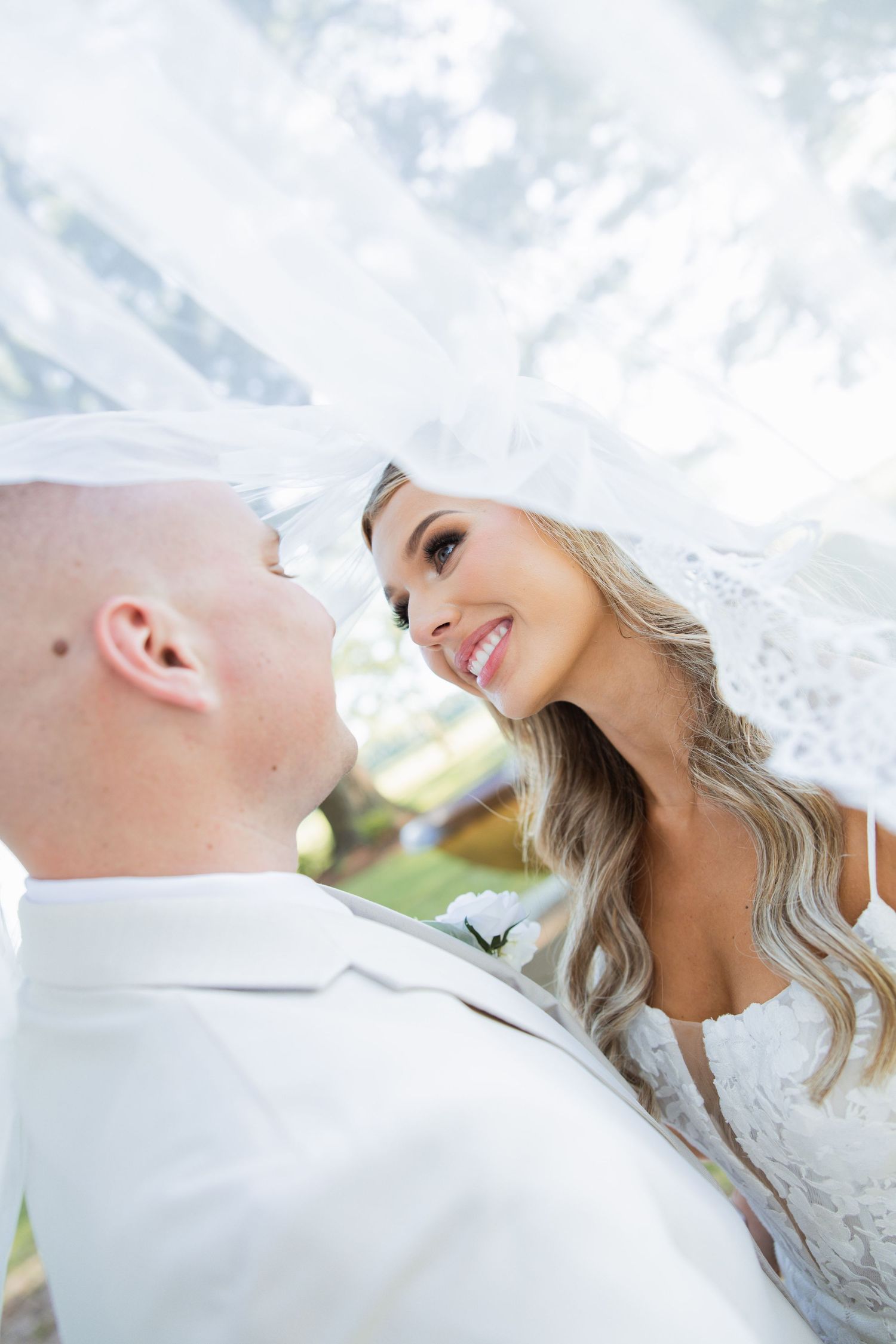 Bride and groom share an intimate moment under a flowing white veil, smiling at each other on their wedding day.