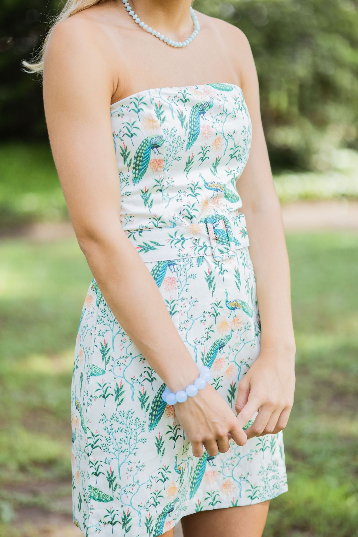 Woman wearing a white strapless floral dress with teal peacock print, blue bracelet and necklace outdoors.