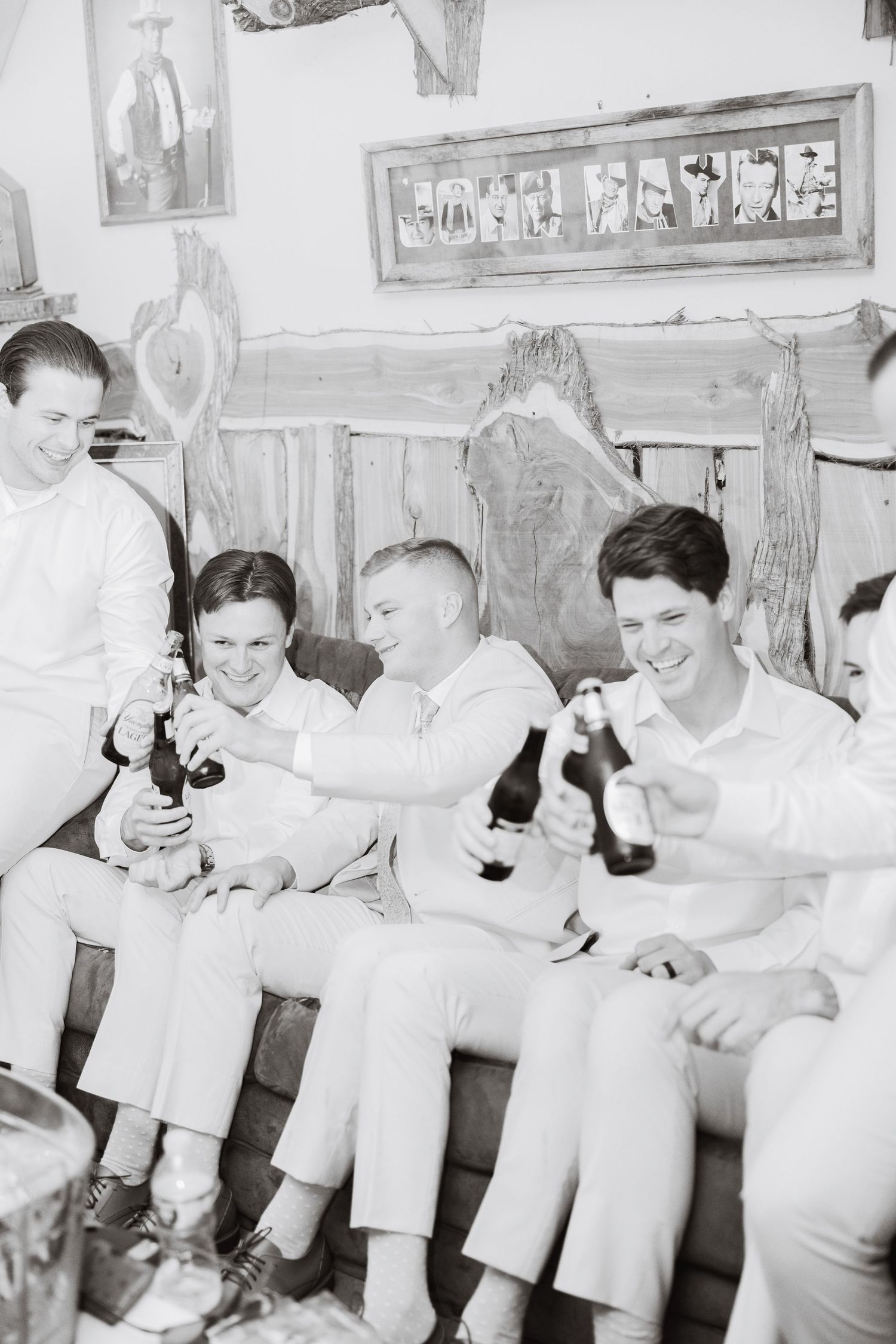 Black and white photo of groom and groomsmen laughing and toasting with beer bottles.