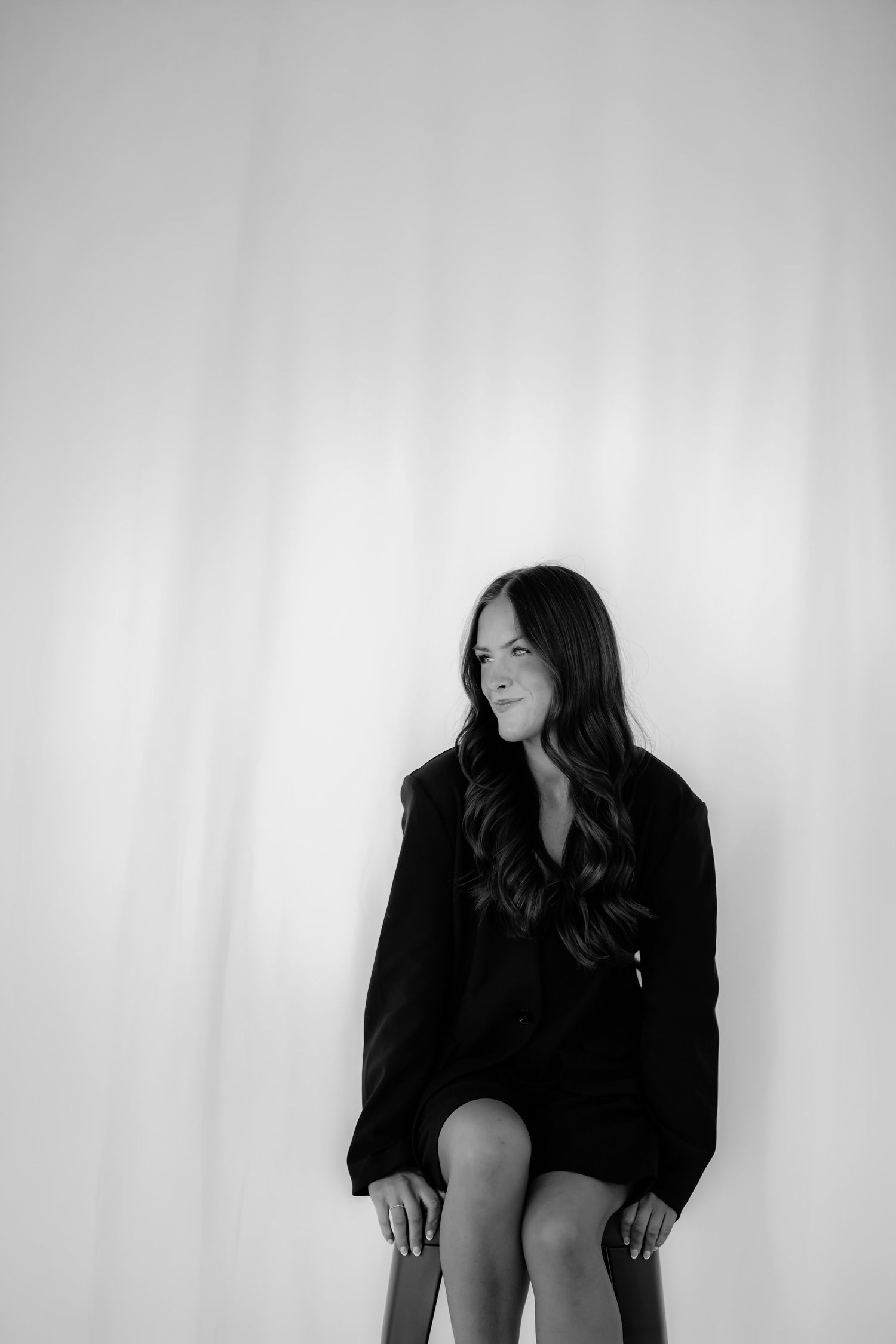 Black and white portrait of a young woman with long dark hair wearing an oversized blazer, seated against a light backdrop.