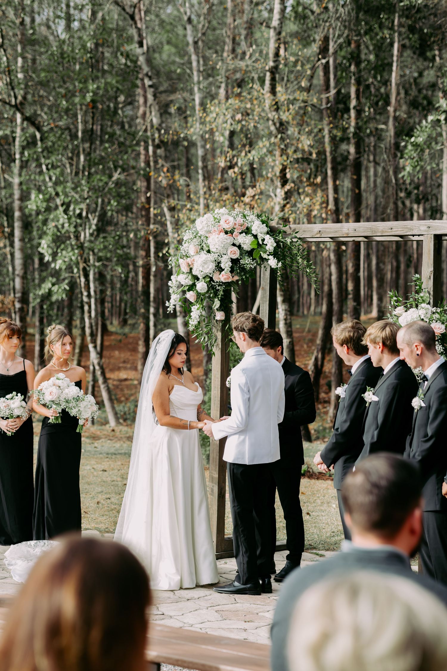 Outdoor forest wedding ceremony with couple at floral arch, bridesmaids in black, groomsmen in black.