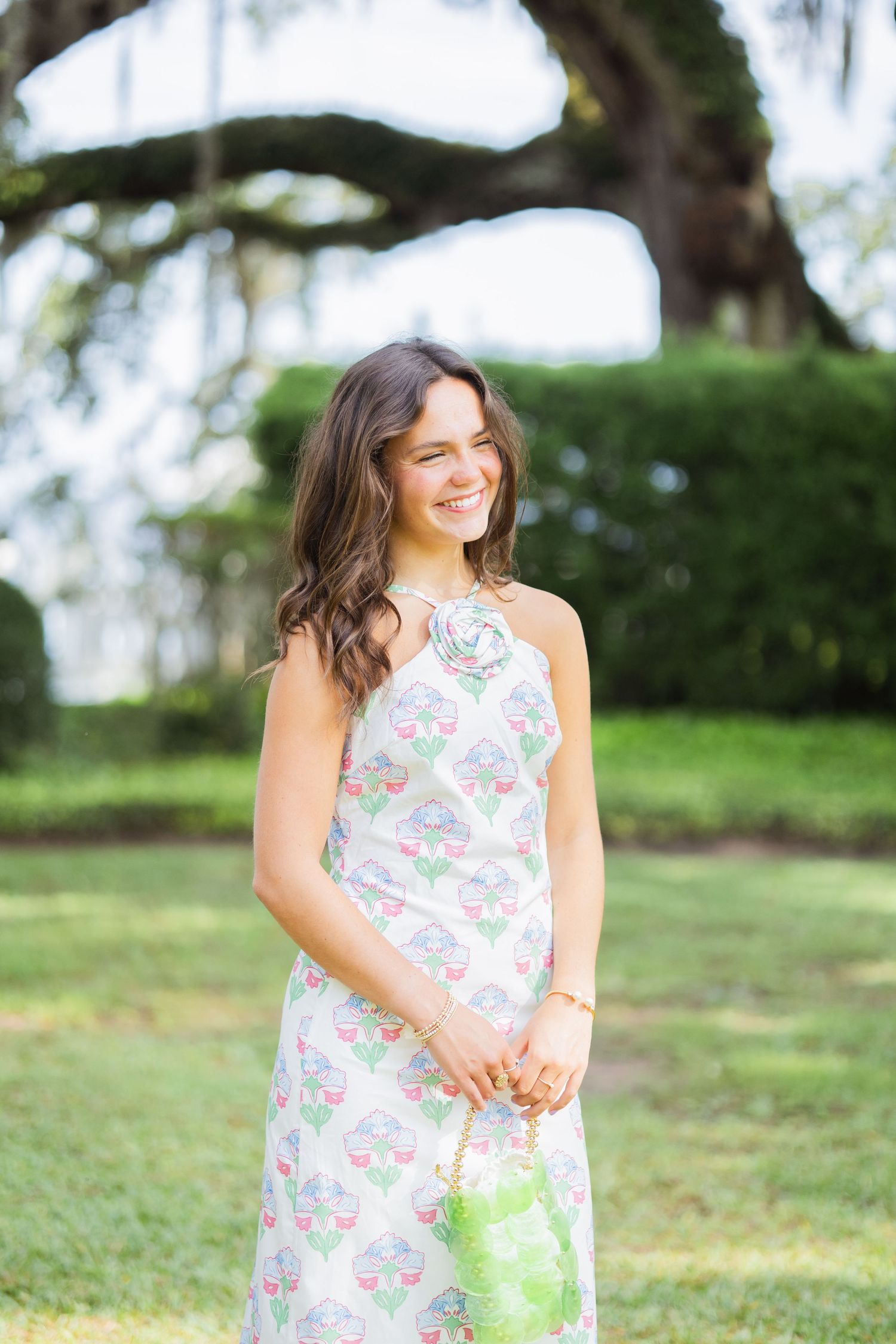 Smiling brunette woman in floral halter maxi dress holding green bag in sunny park.