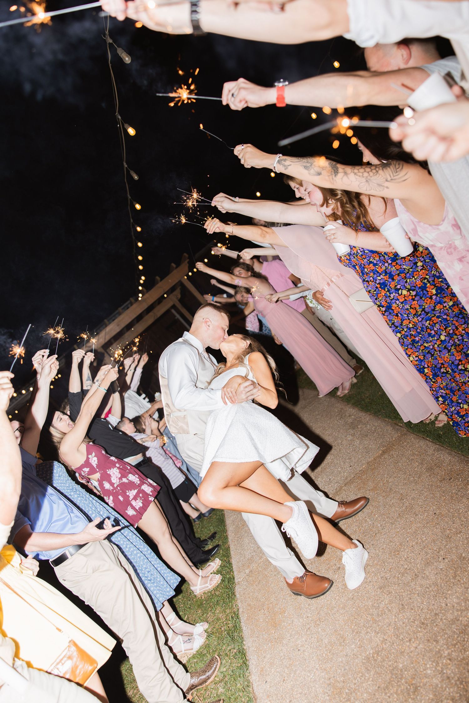 Bride and groom kiss during sparkler sendoff lined with cheering wedding guests at night.