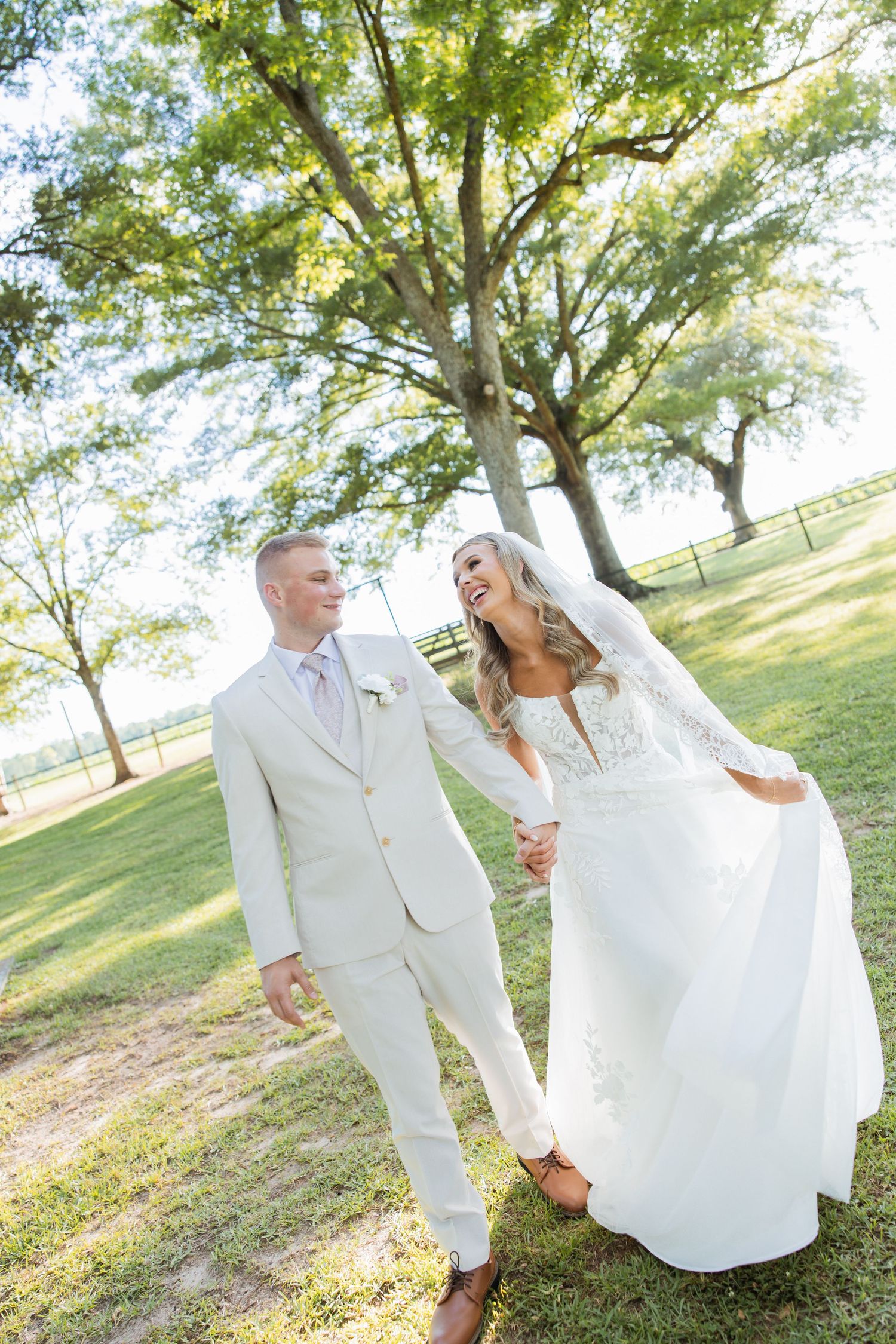 Smiling bride and groom walk hand in hand through park on sunny wedding day.