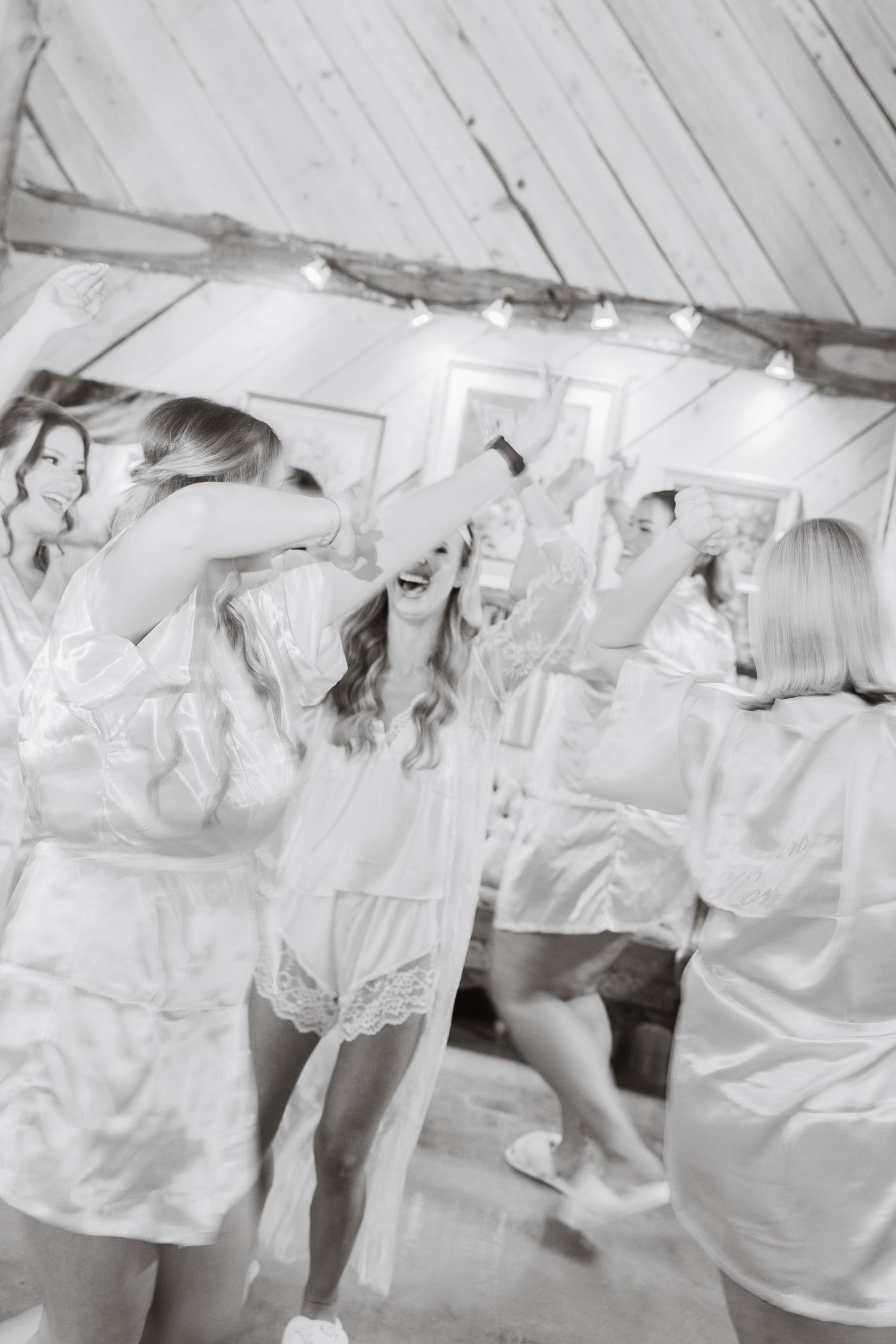 Black and white photo of bridesmaids in white robes celebrating and dancing together before the wedding.