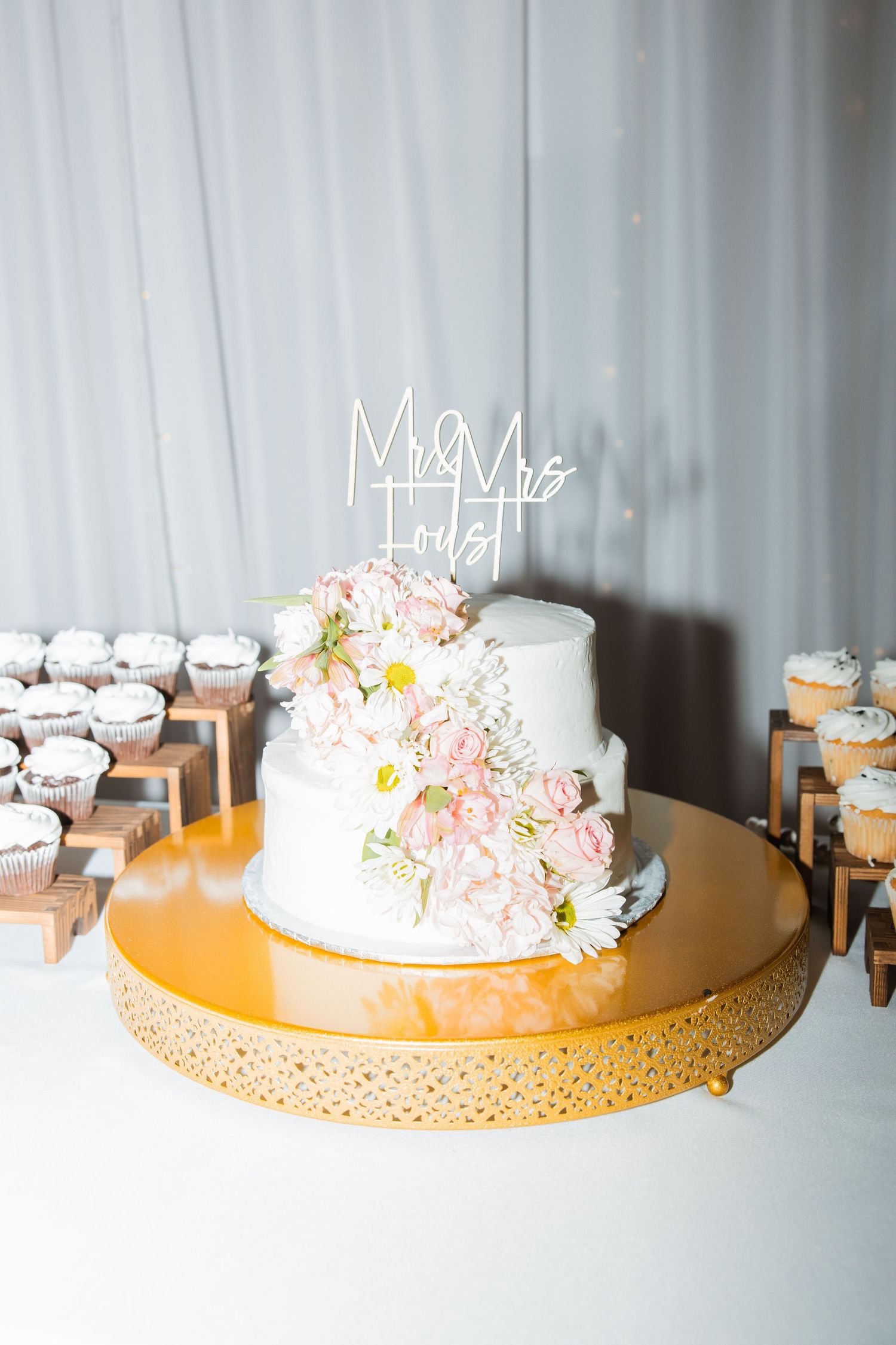 Two-tier white wedding cake with pink roses and daisies on a gold stand with Mr and Mrs topper.