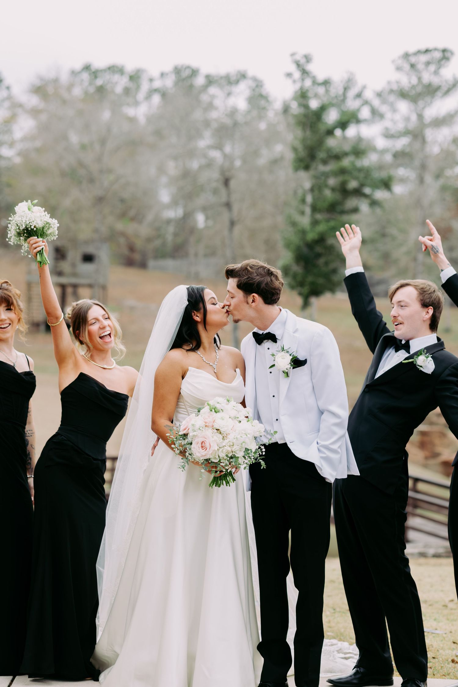 Joyful wedding party celebrating as bride and groom kiss, bridesmaids in black gowns raising bouquets.