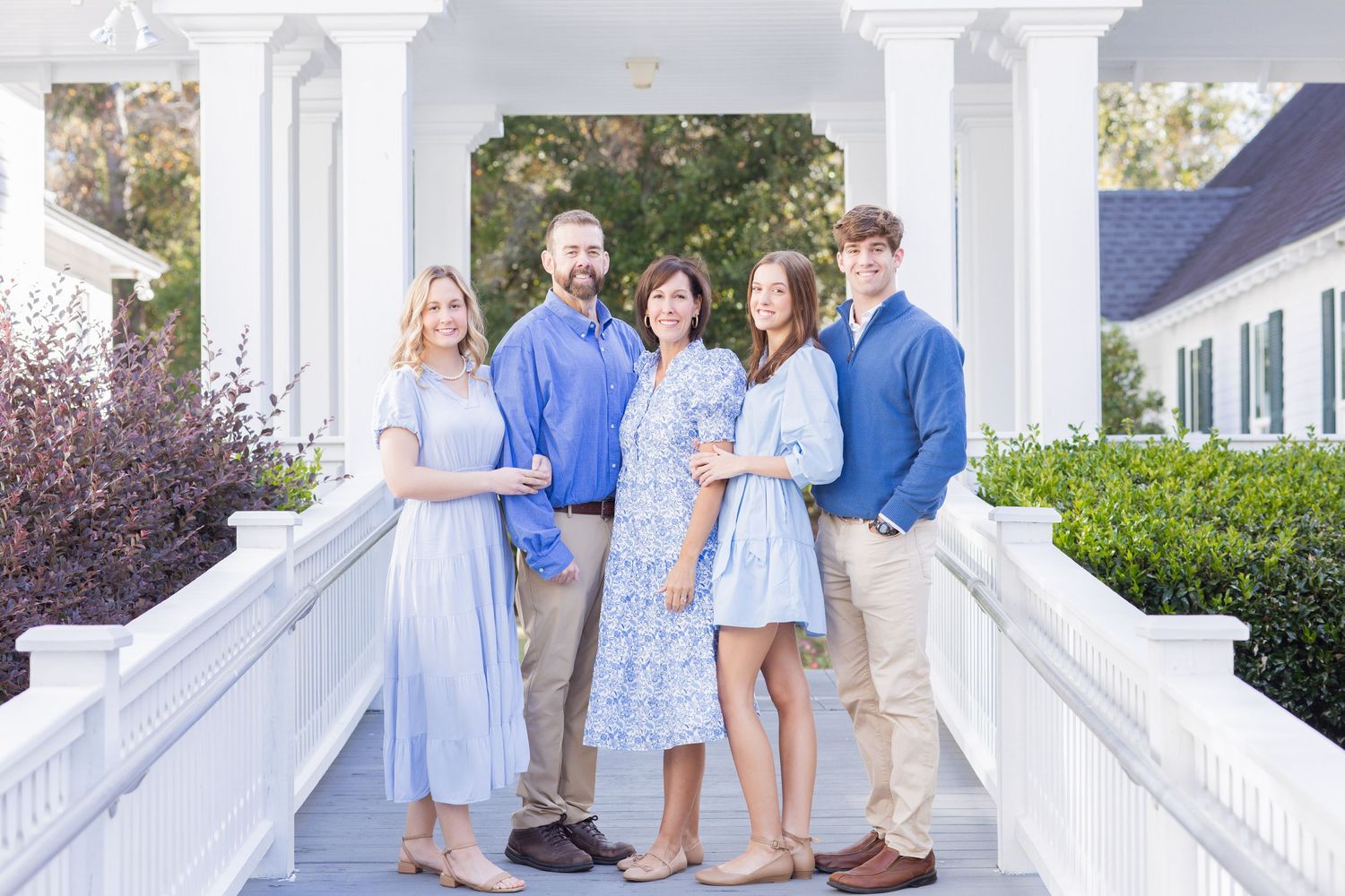 Family of five smiling together on a white columned porch in coordinating blue and tan outfits.