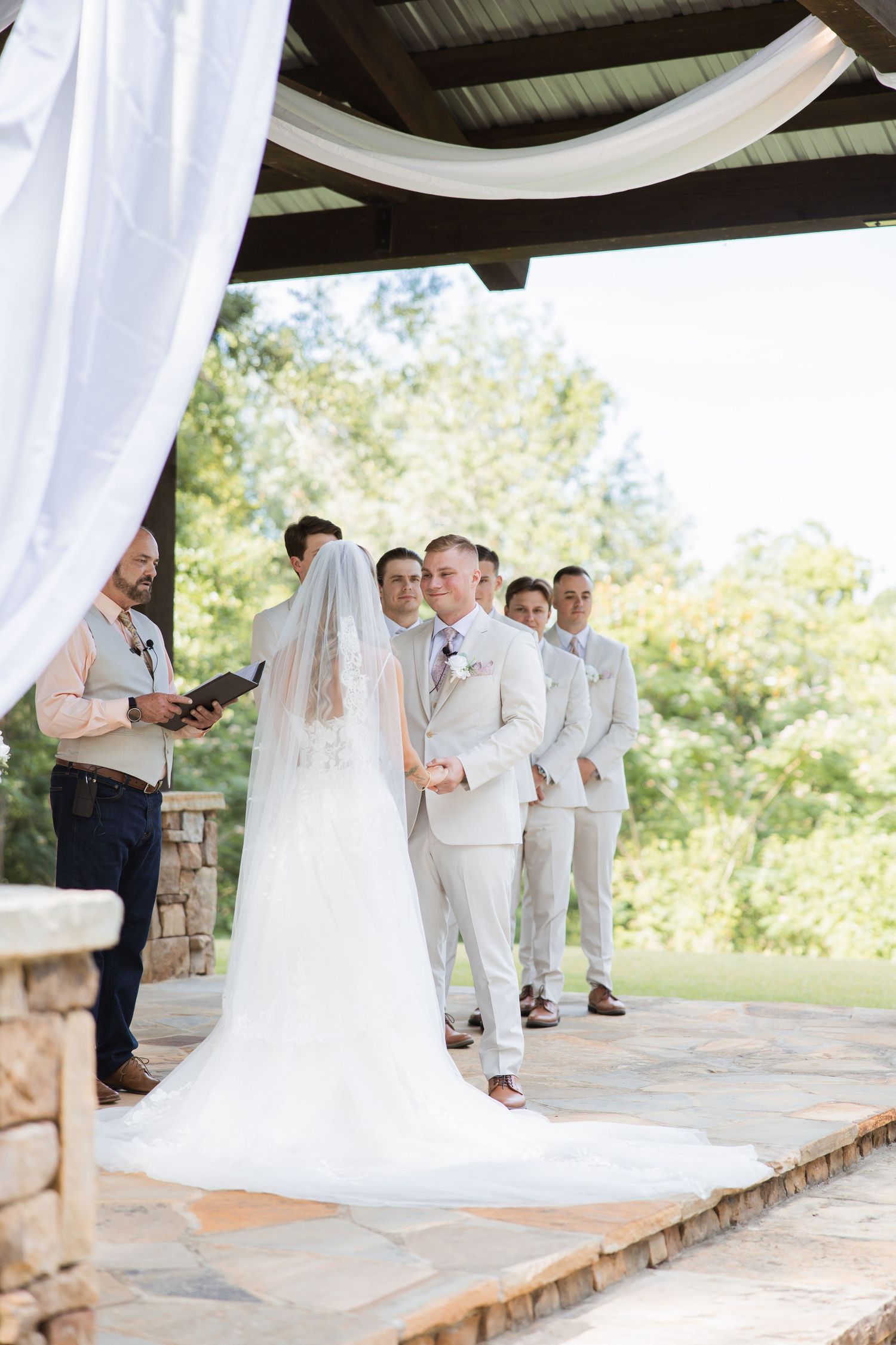 Bride and groom exchange vows at outdoor ceremony under pergola with groomsmen standing nearby.