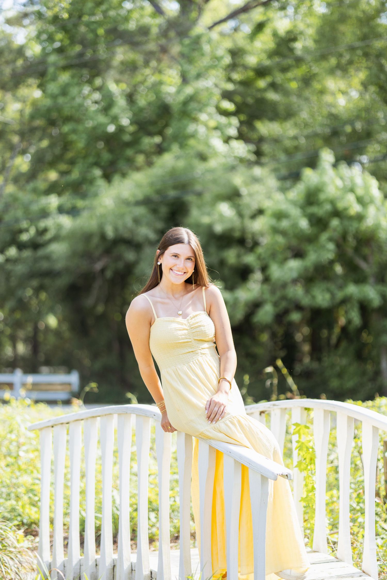 Young woman in yellow maxi dress leaning on white bridge railing surrounded by lush green trees.