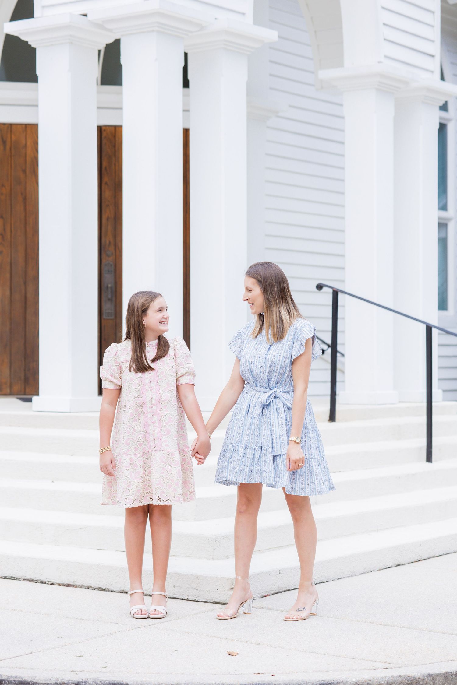 A mother and daughter hold hands and smile at each other in front of a white building with tall columns.