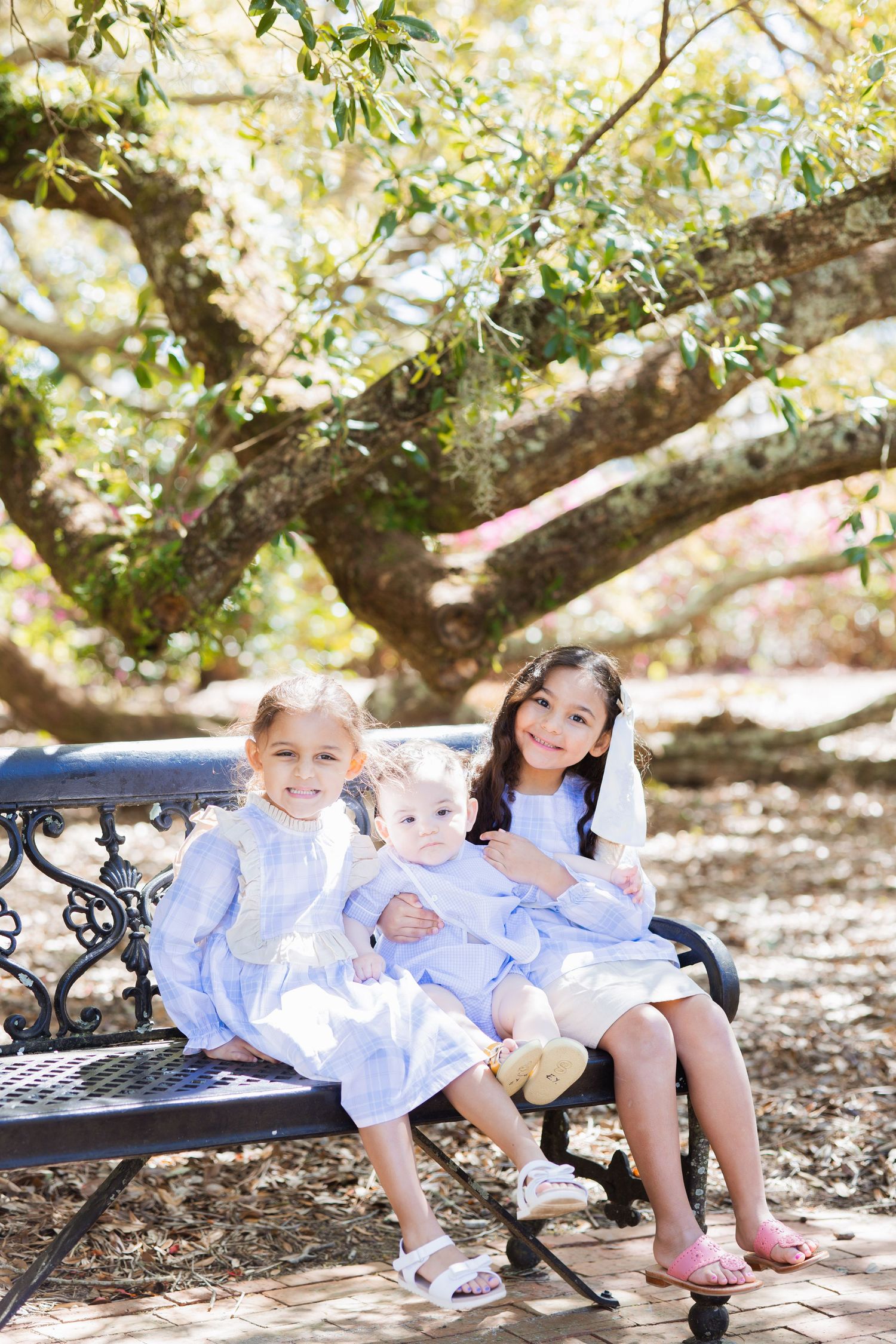 Three young sisters in matching blue dresses sitting on an ornate iron bench outdoors under a large tree.