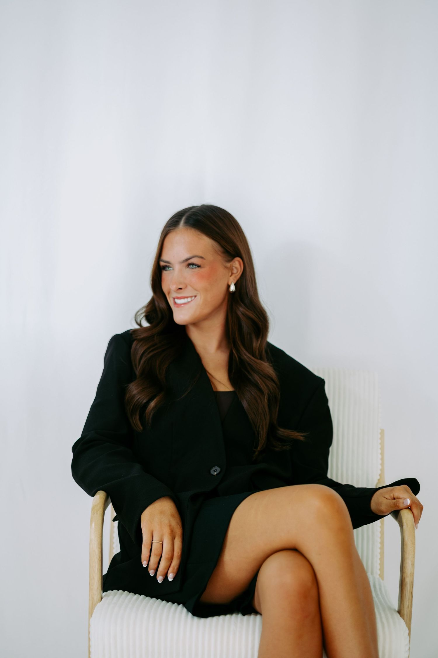 Smiling brunette woman in black blazer and dress seated in white chair against light background.