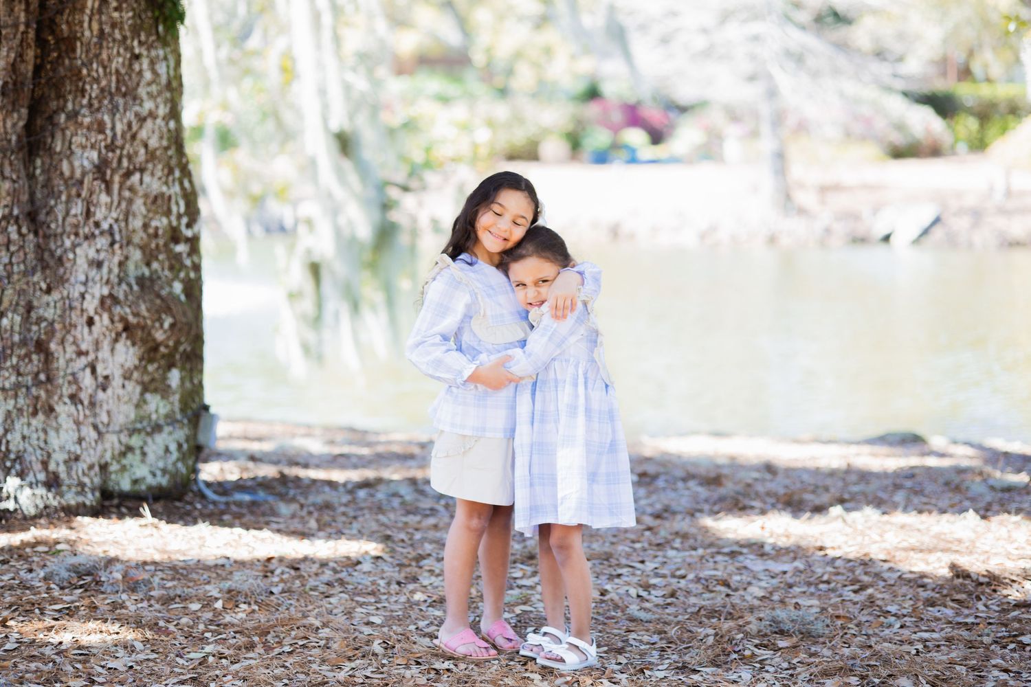 Two young sisters in blue dresses hugging each other near a tree by a lake in a park.