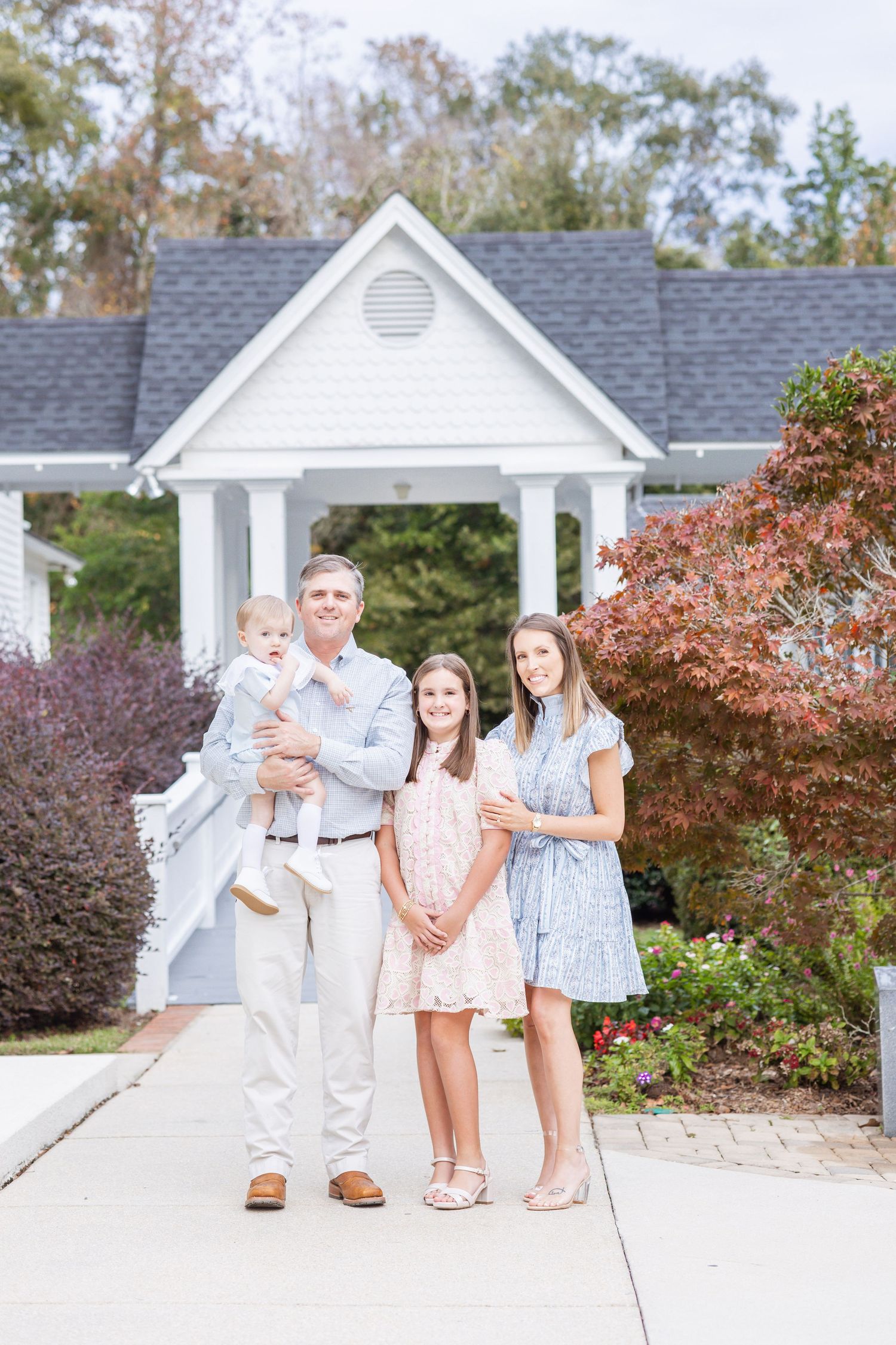 A father holds a baby while posing with two daughters in front of a white gazebo surrounded by colorful foliage.