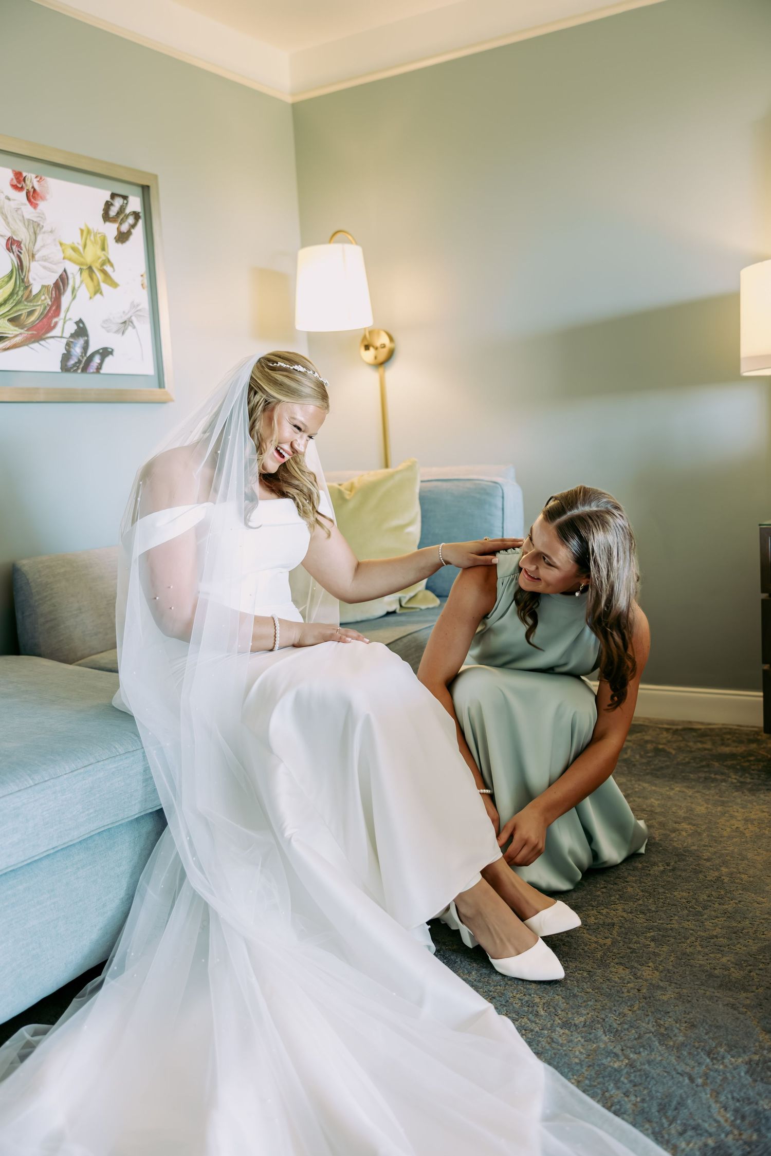 Bridesmaid helping smiling bride put on white heels while seated in an elegantly decorated hotel suite.
