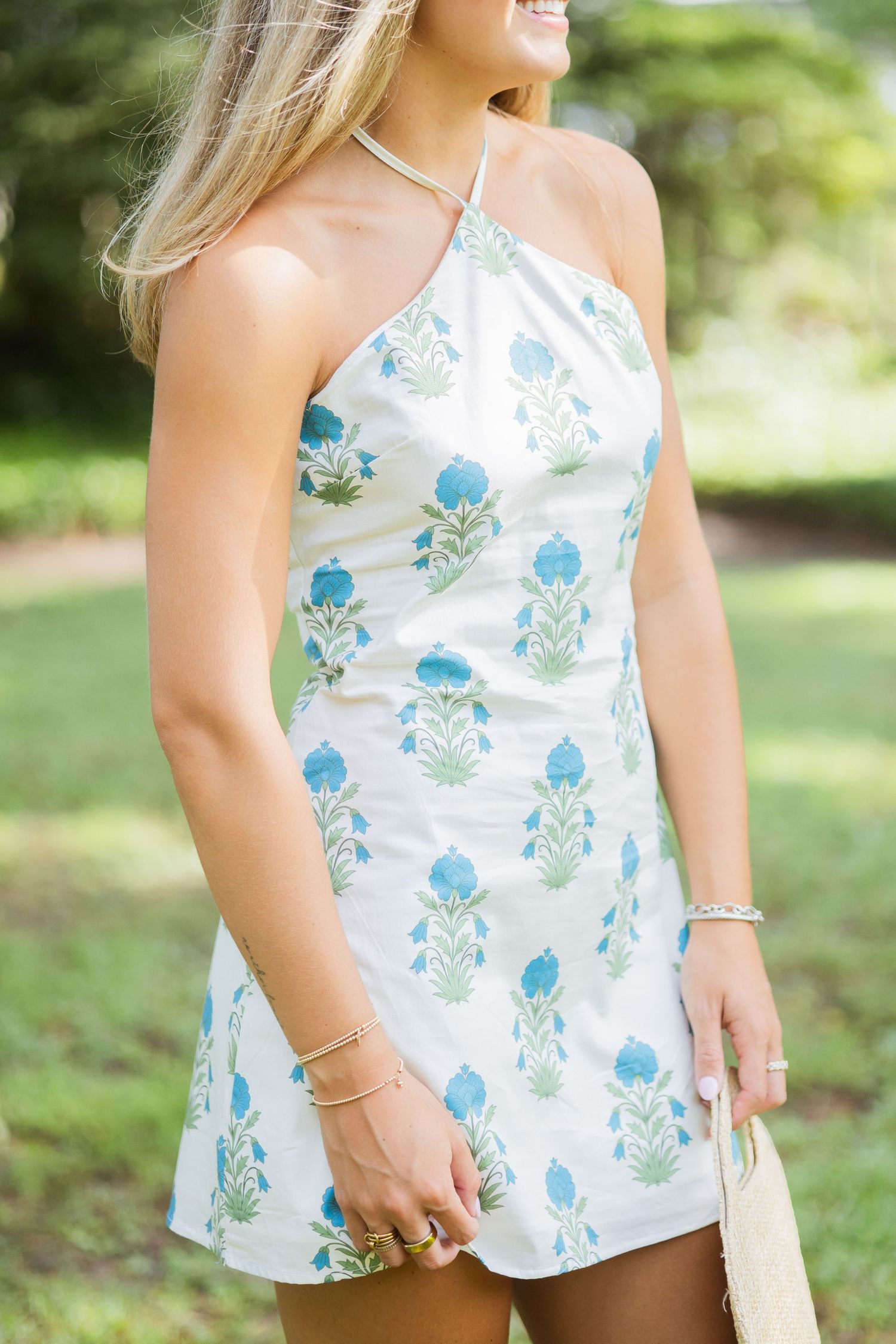 Blonde woman in white and blue floral halter mini dress holding straw bag outdoors in garden.