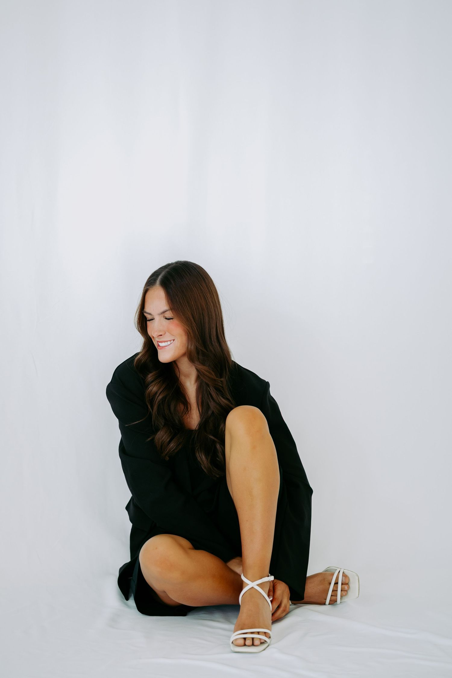 Smiling brunette woman in black dress seated on floor adjusting white strappy heeled sandals.