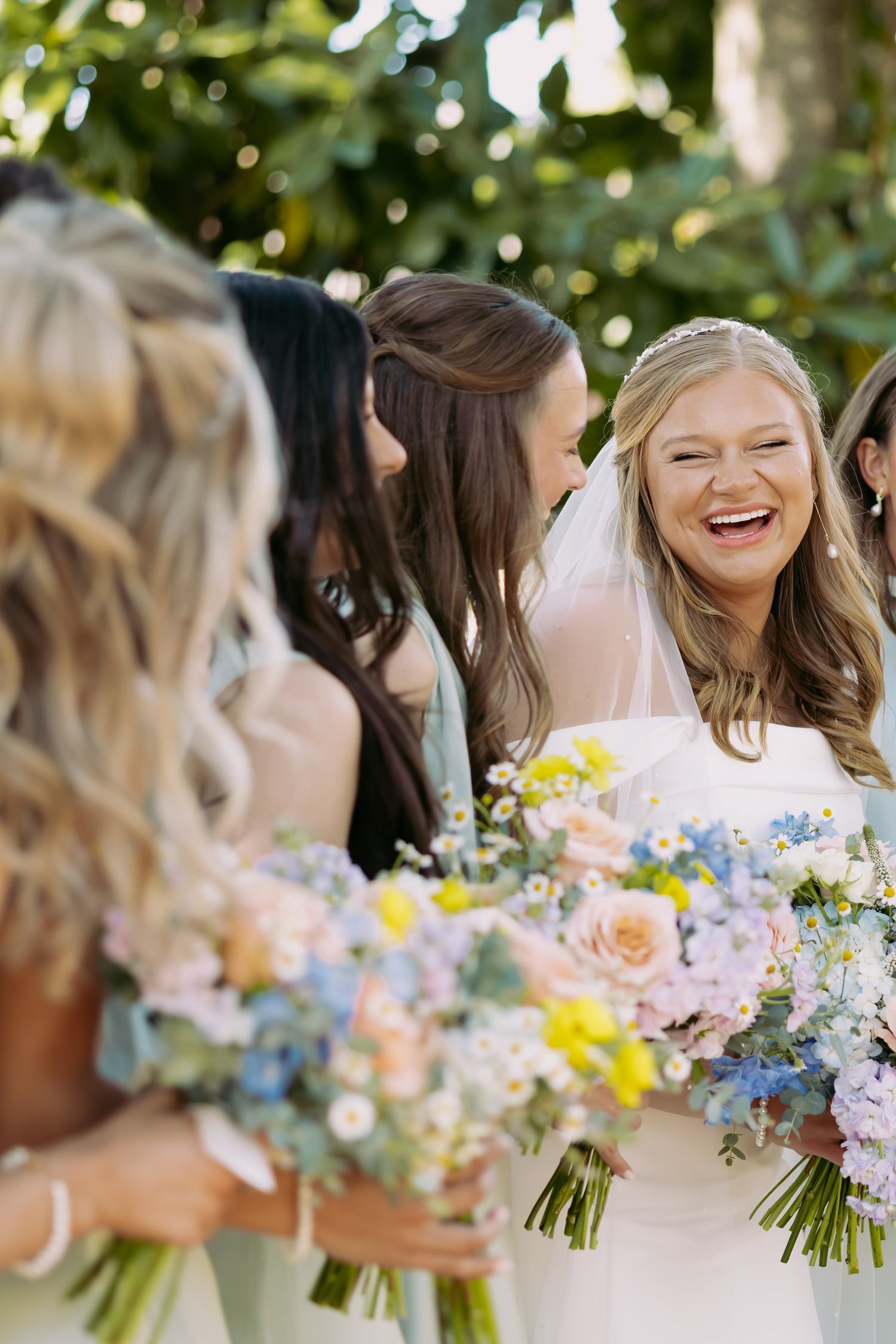 Smiling bride in white gown holding colorful bouquet surrounded by laughing bridesmaids outdoors.