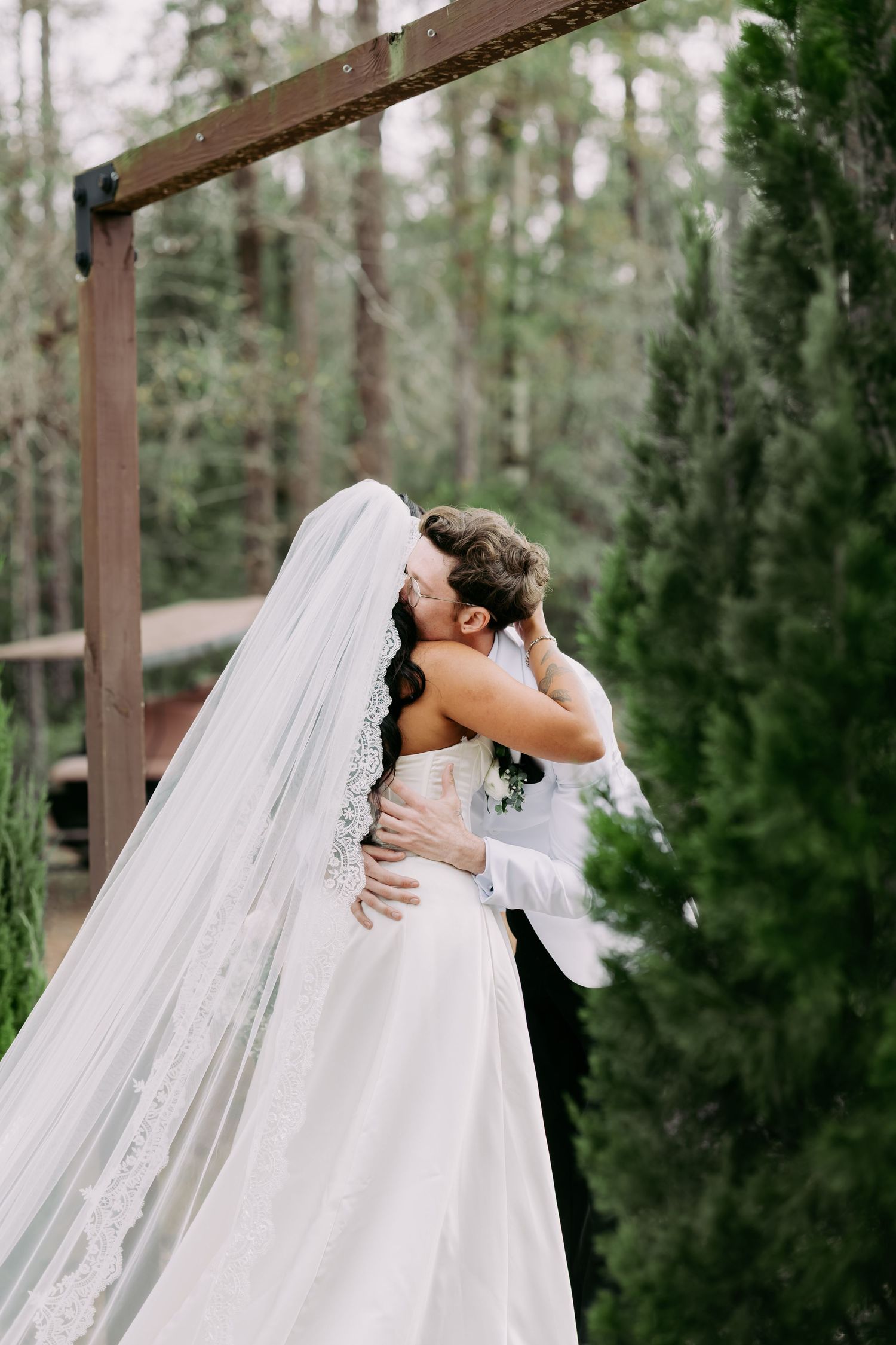 Bride and groom embrace near wooden arch surrounded by tall evergreen trees, her cathedral lace veil trailing behind.