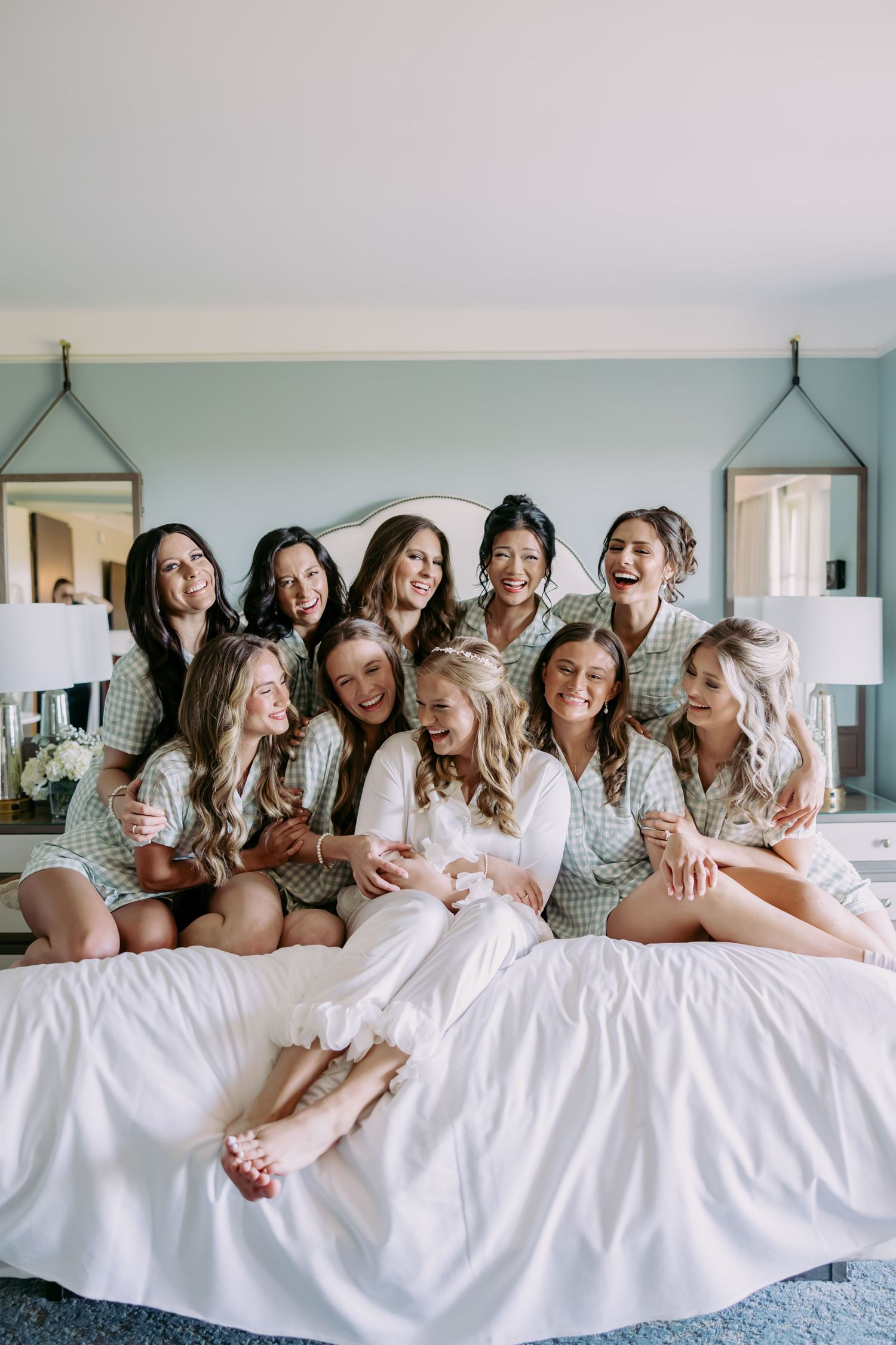 Bride in white robe surrounded by bridesmaids in matching green plaid pajamas laughing together on a bed.