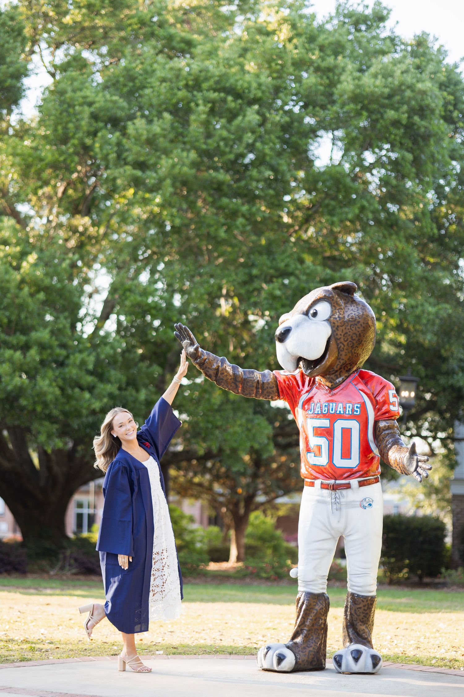 Female graduate in cap and gown high-fiving Jaguars mascot statue on sunny campus grounds.