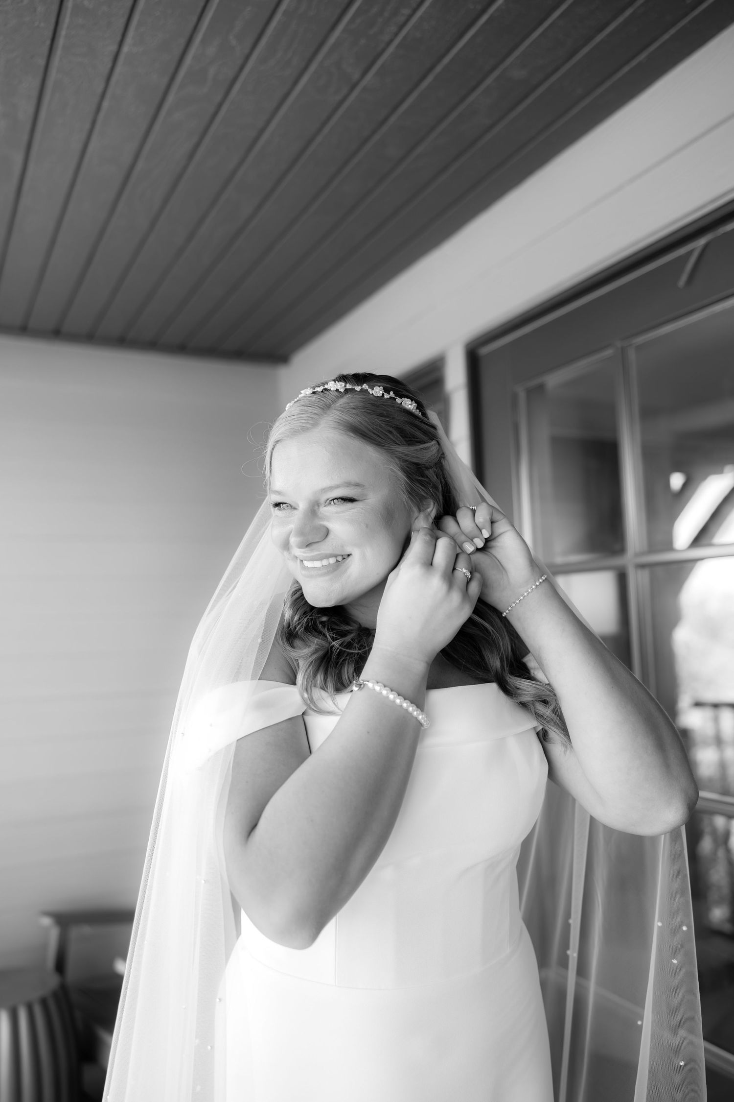 Smiling bride in white gown and veil adjusting her earrings in a black and white wedding portrait.