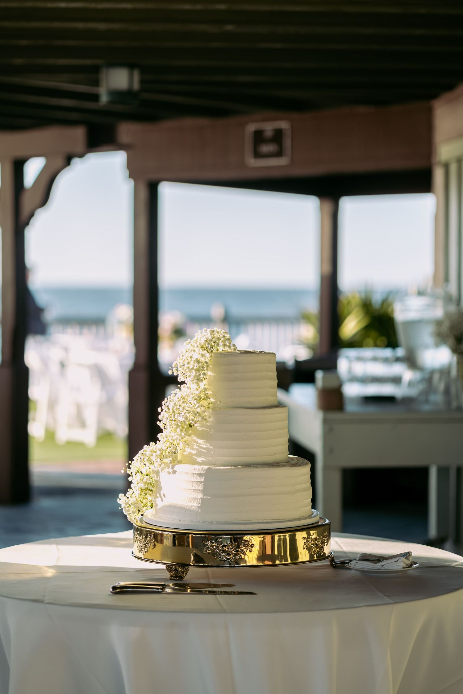 Elegant four-tier white wedding cake with baby's breath flowers on a gold stand, oceanfront venue backdrop.
