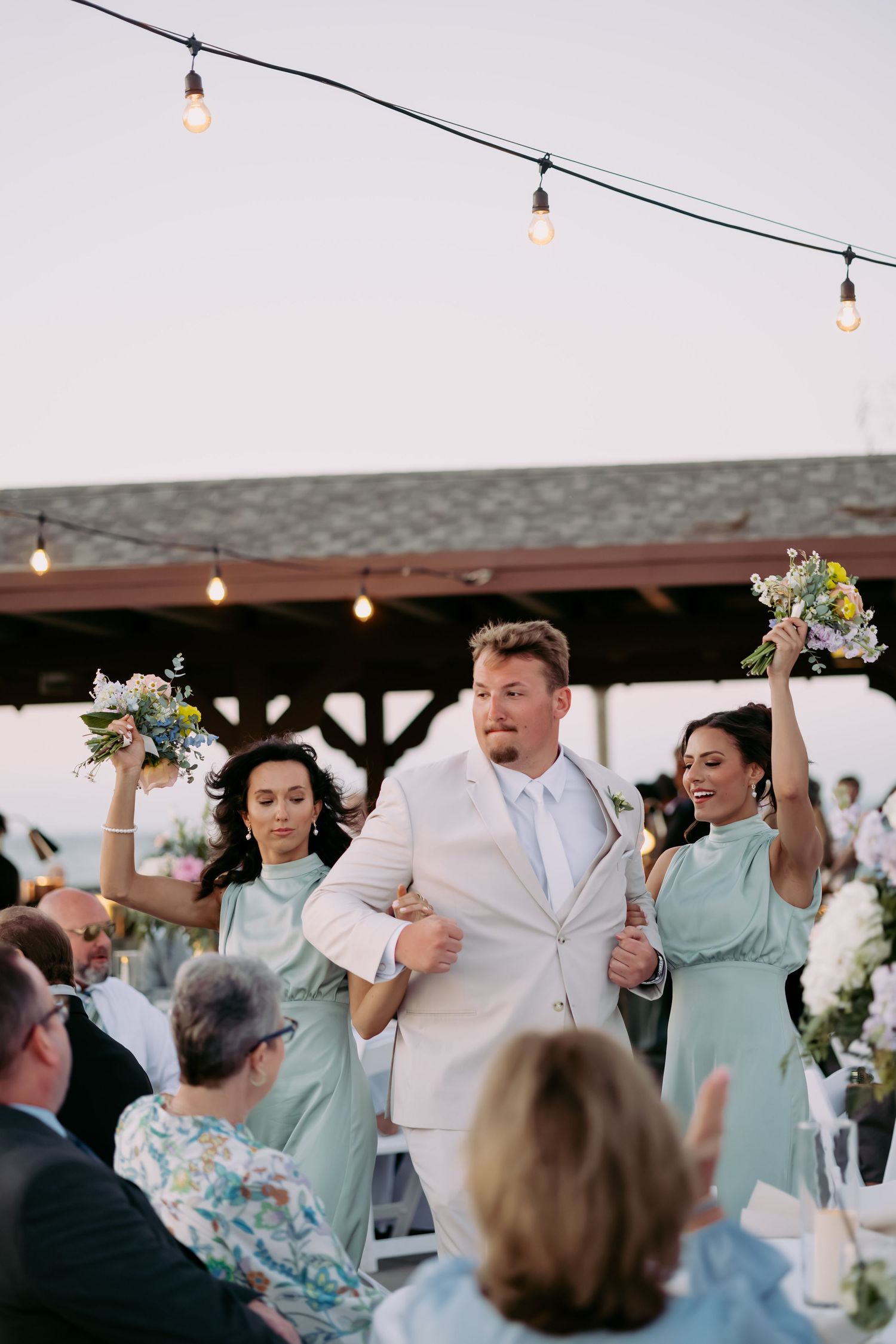 Groom in cream suit walks with bridesmaids in mint dresses holding bouquets aloft under string lights at outdoor reception.
