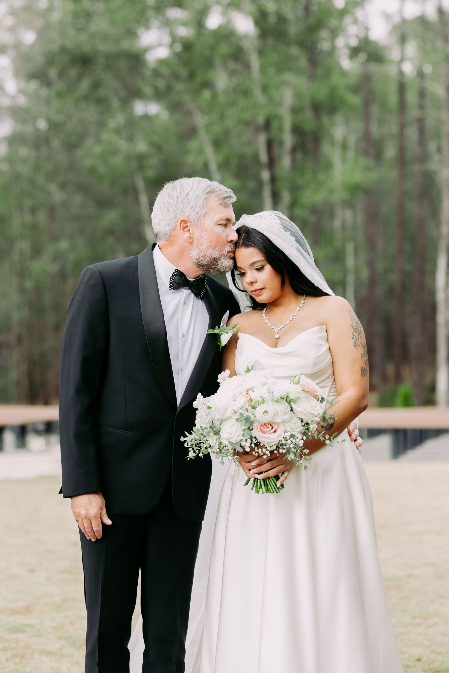 Groom in black tuxedo tenderly kissing bride's forehead as she holds a blush bouquet outdoors.