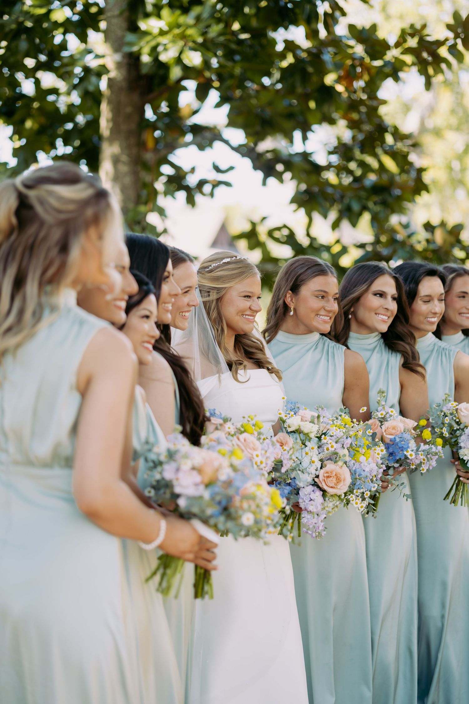 Bride in white gown stands with bridesmaids in mint green dresses holding colorful spring bouquets.