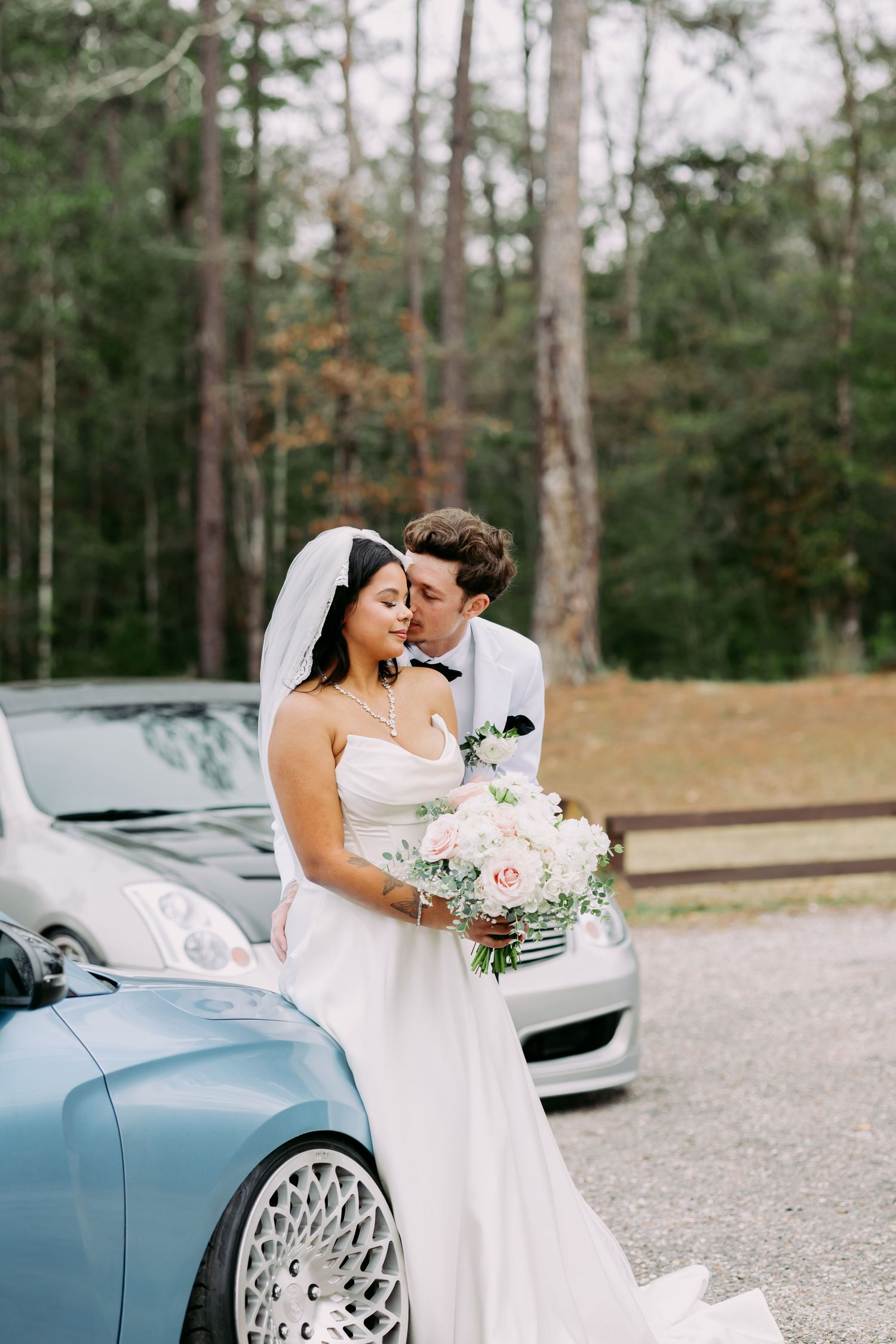 Bride and groom embrace tenderly beside a baby blue custom car in a forest setting.