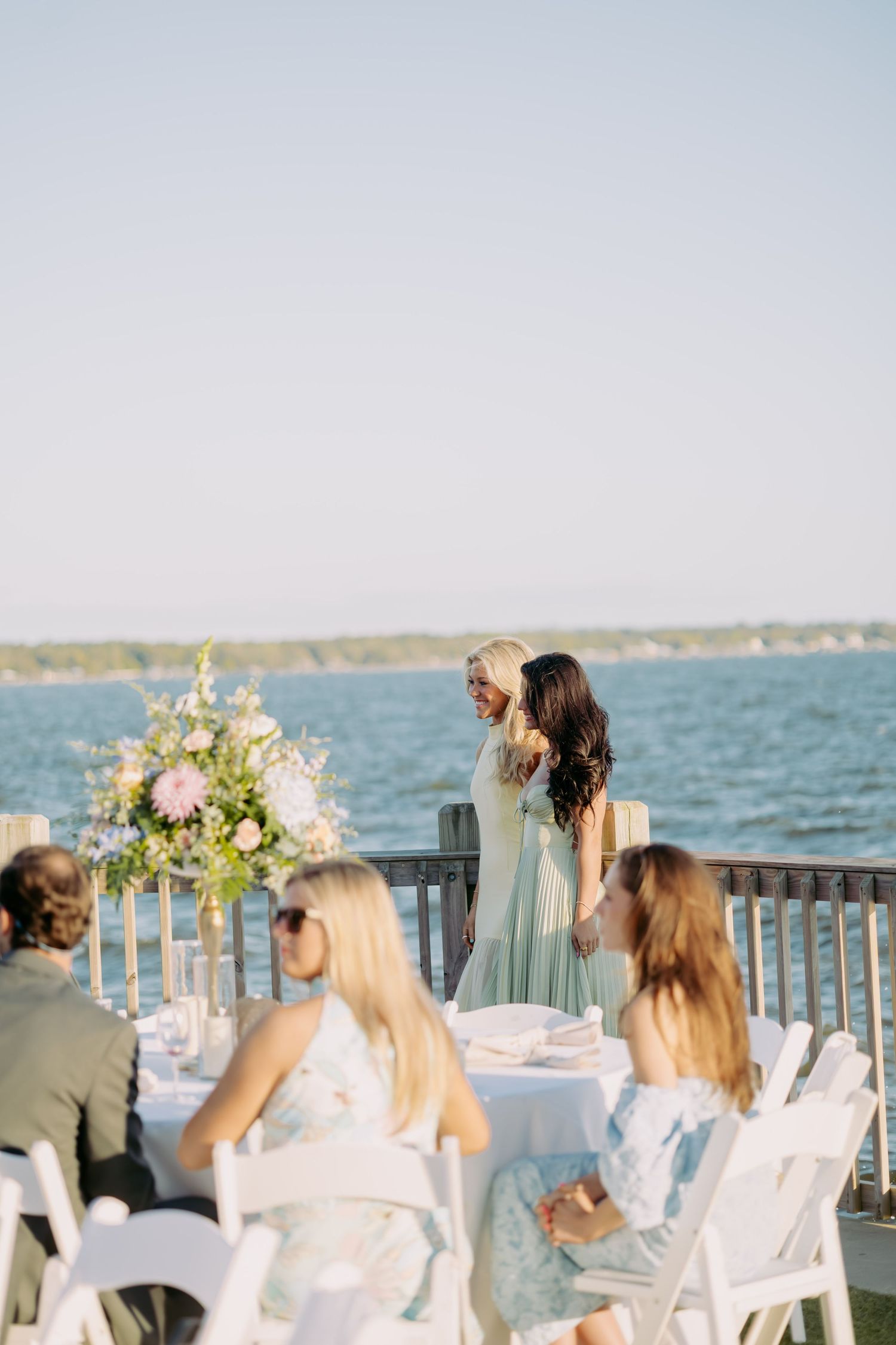 Bridesmaids in sage green dresses chatting near floral centerpiece at elegant waterfront wedding reception.