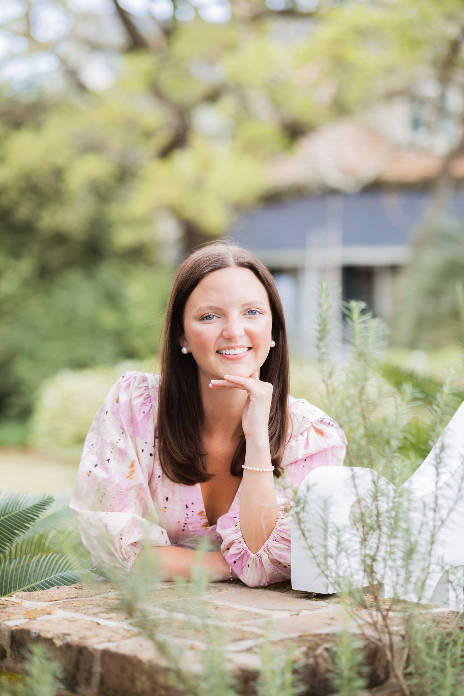 Smiling brunette woman in a pink floral dress resting her chin on her hand in a garden.