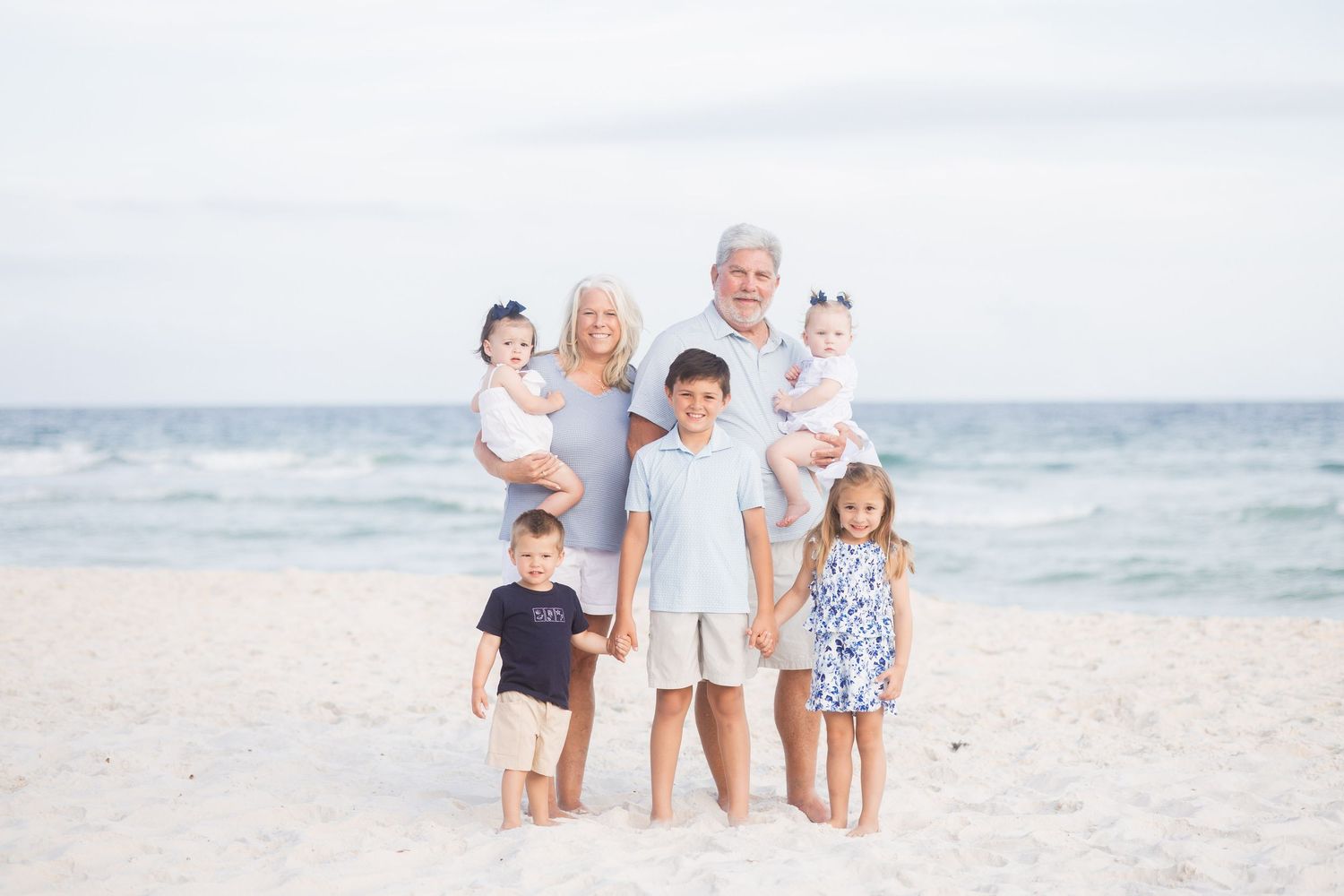 Grandparents with five young grandchildren smiling together on a white sandy beach at sunset.