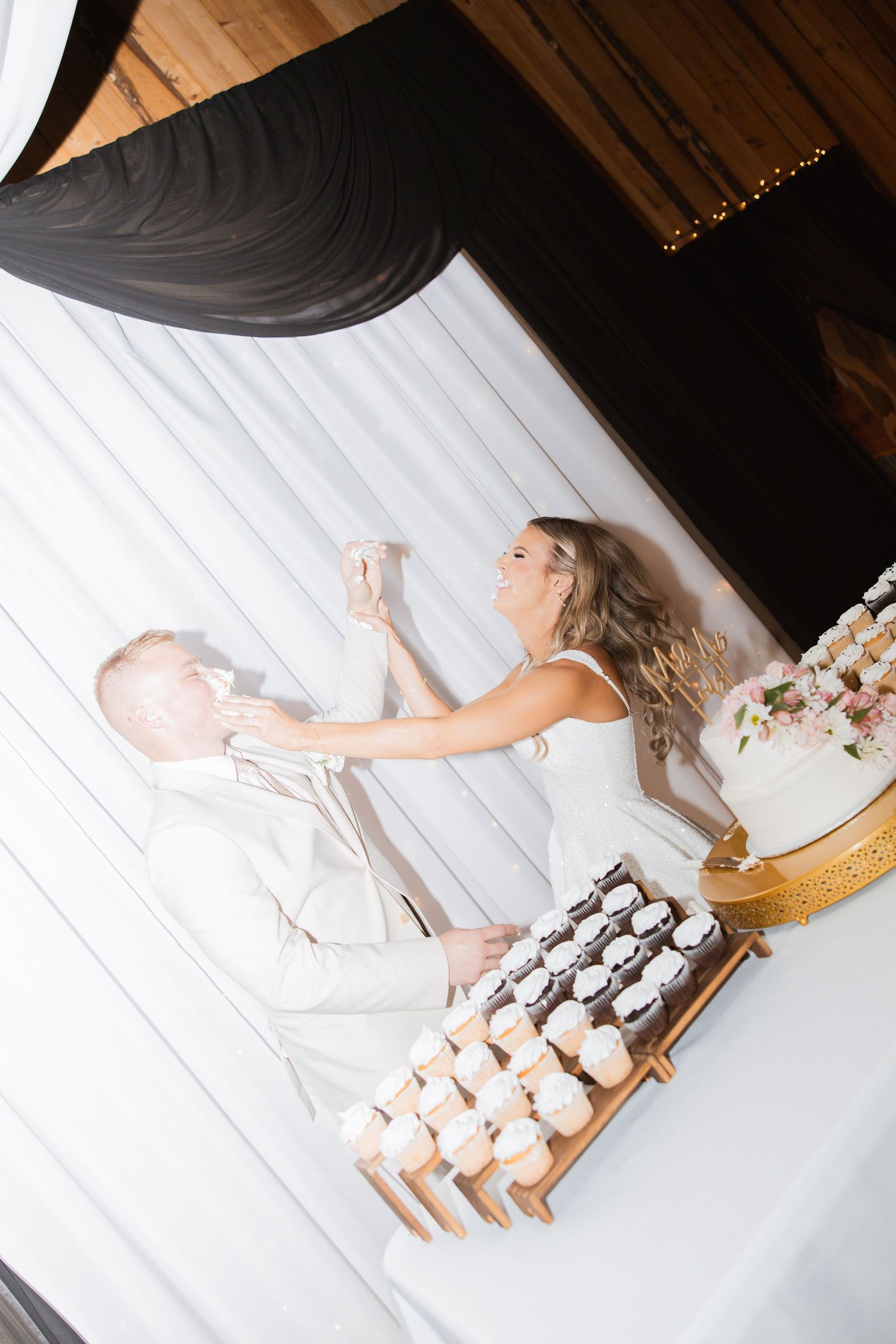 Bride laughs while feeding wedding cake to groom at elegant reception with cupcake display.