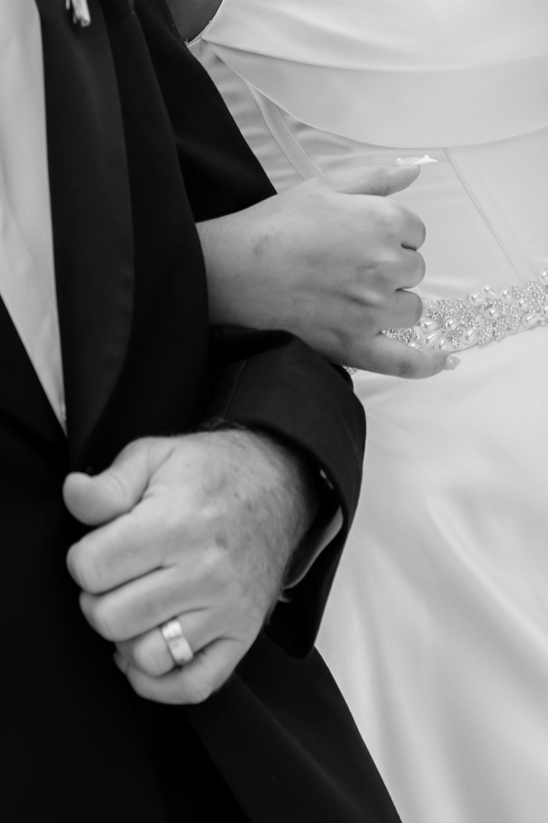 Close-up black and white photo of wedding couple holding hands, groom wearing a ring and black tuxedo.