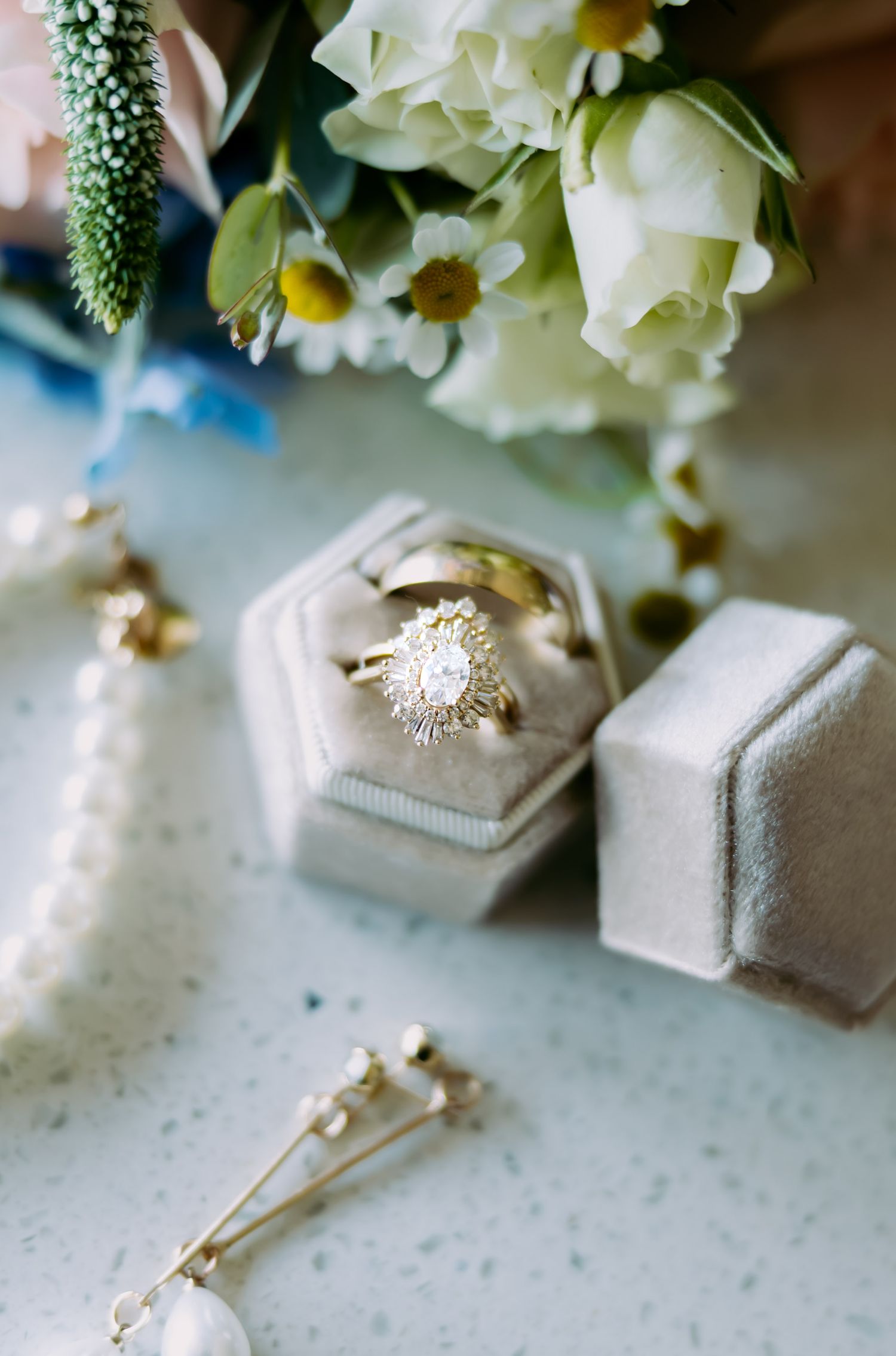 Gold diamond halo engagement ring displayed in a velvet hexagonal ring box surrounded by wedding jewelry and white flowers.