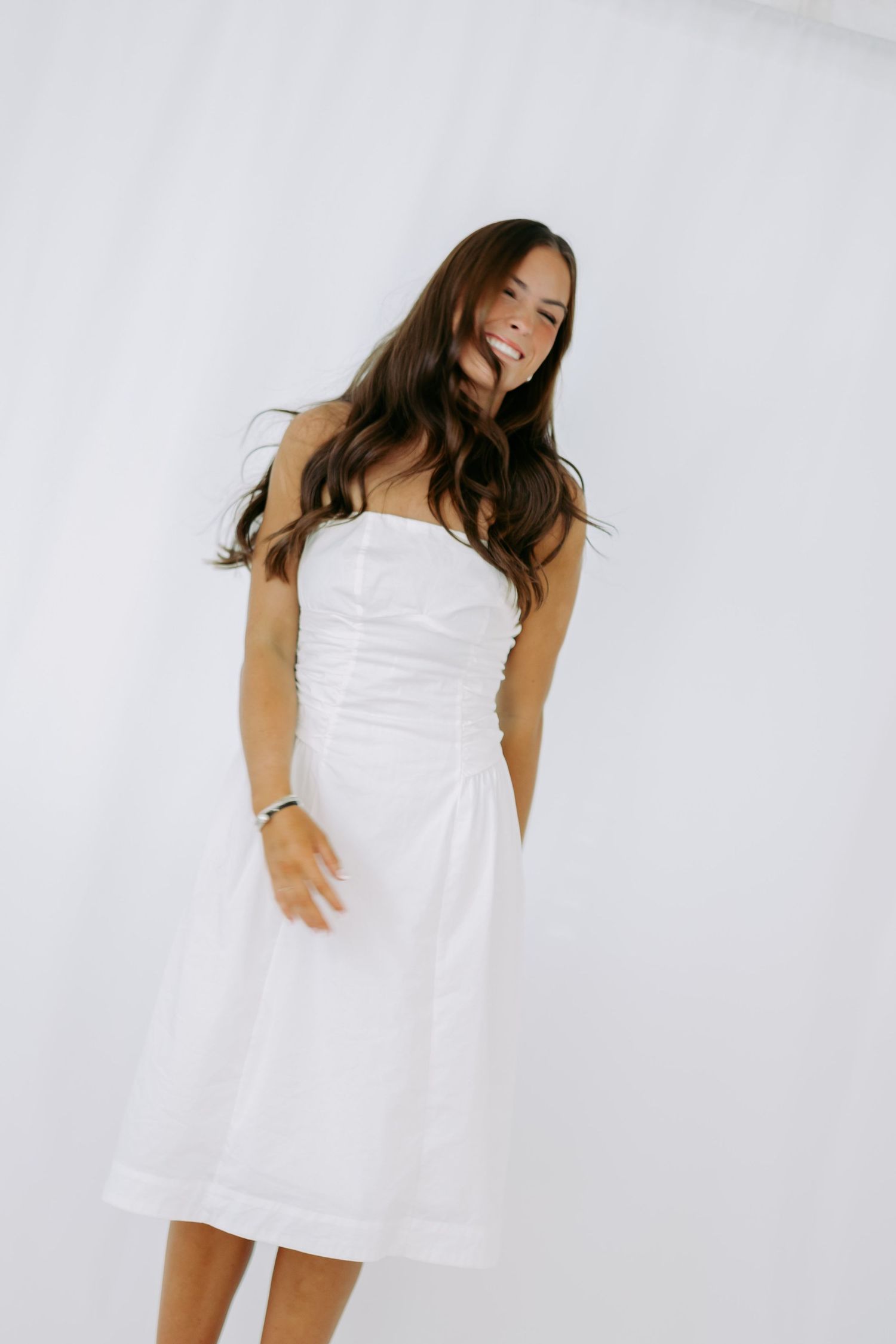 Brunette woman in white strapless ruched midi dress posing dynamically against white background.