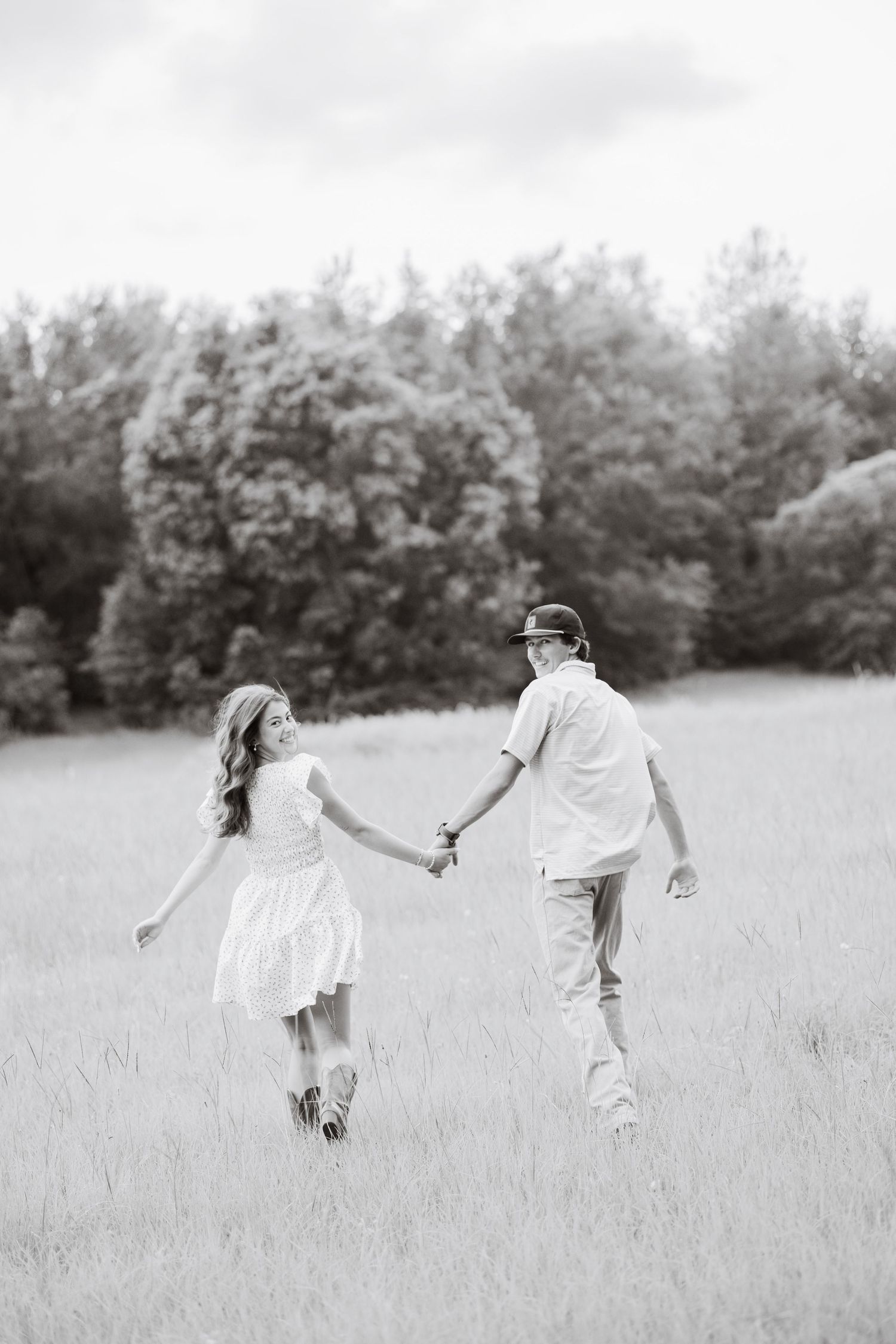 Black and white photo of a couple holding hands and walking through an open grassy field near trees.