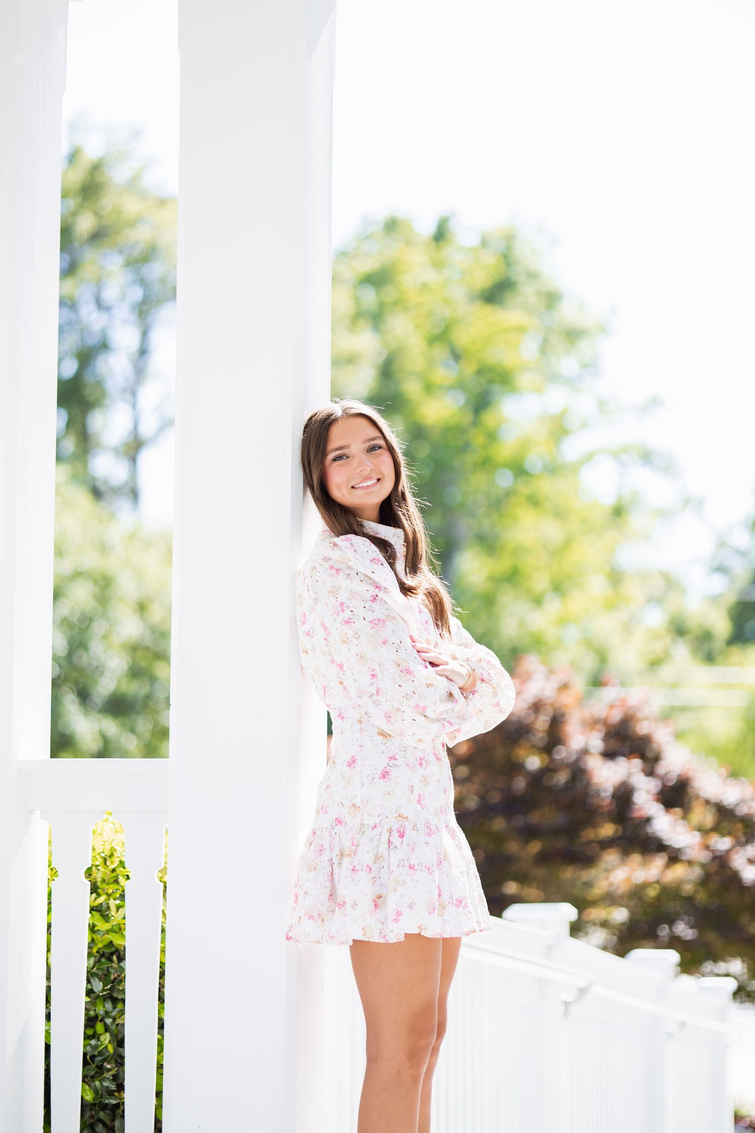 Young woman in floral dress smiling on white porch with columns and green trees in background.