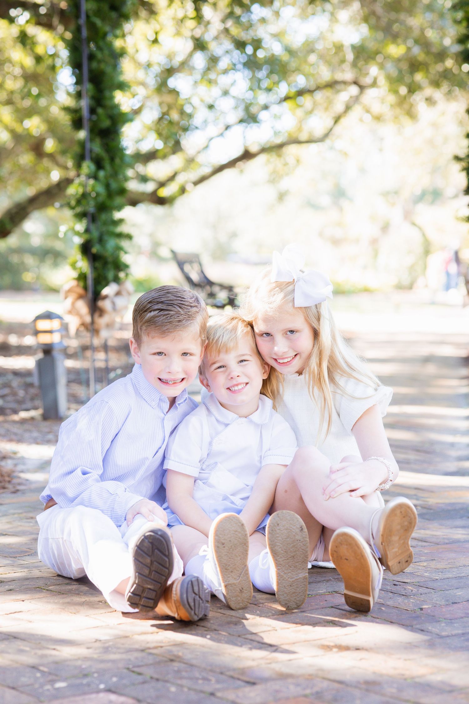 Three young siblings sit together on a brick path, smiling, dressed in white and pastel outfits in a sunny park.