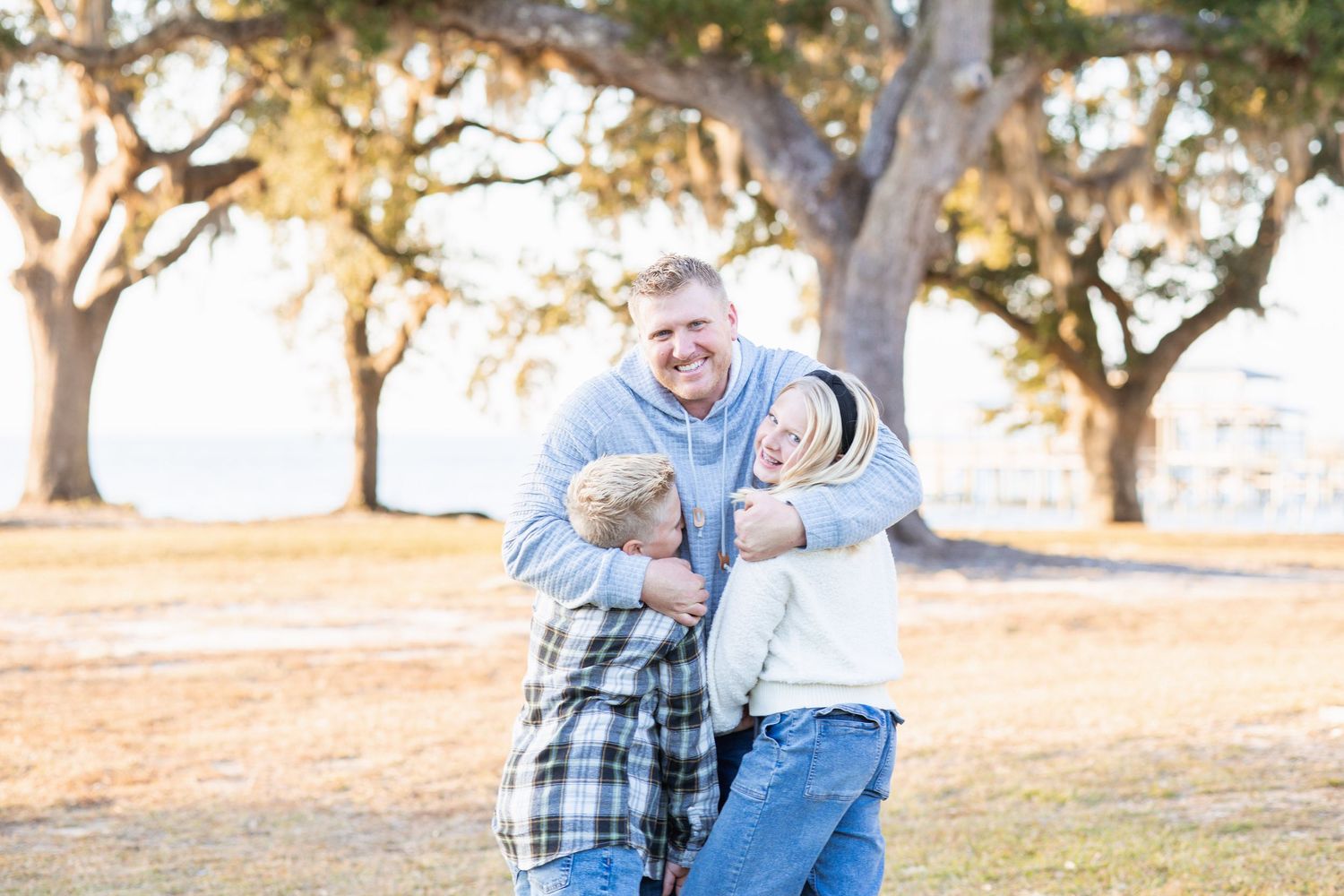 Father laughing while his two children playfully hug him outdoors under large oak trees at golden hour.