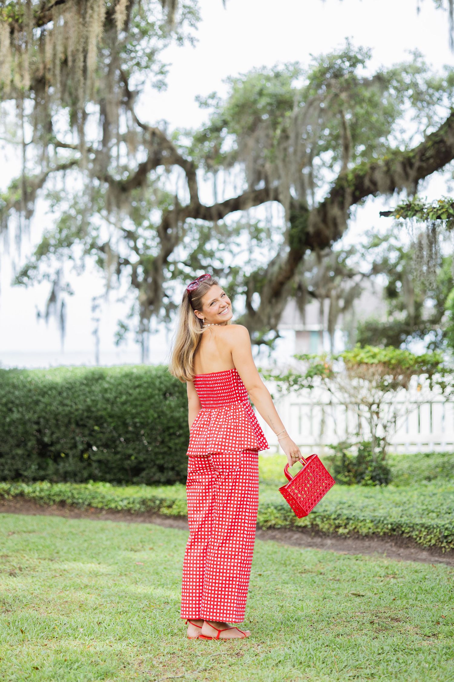 Blonde woman in red polka dot outfit holding red bag under Spanish moss oak tree.