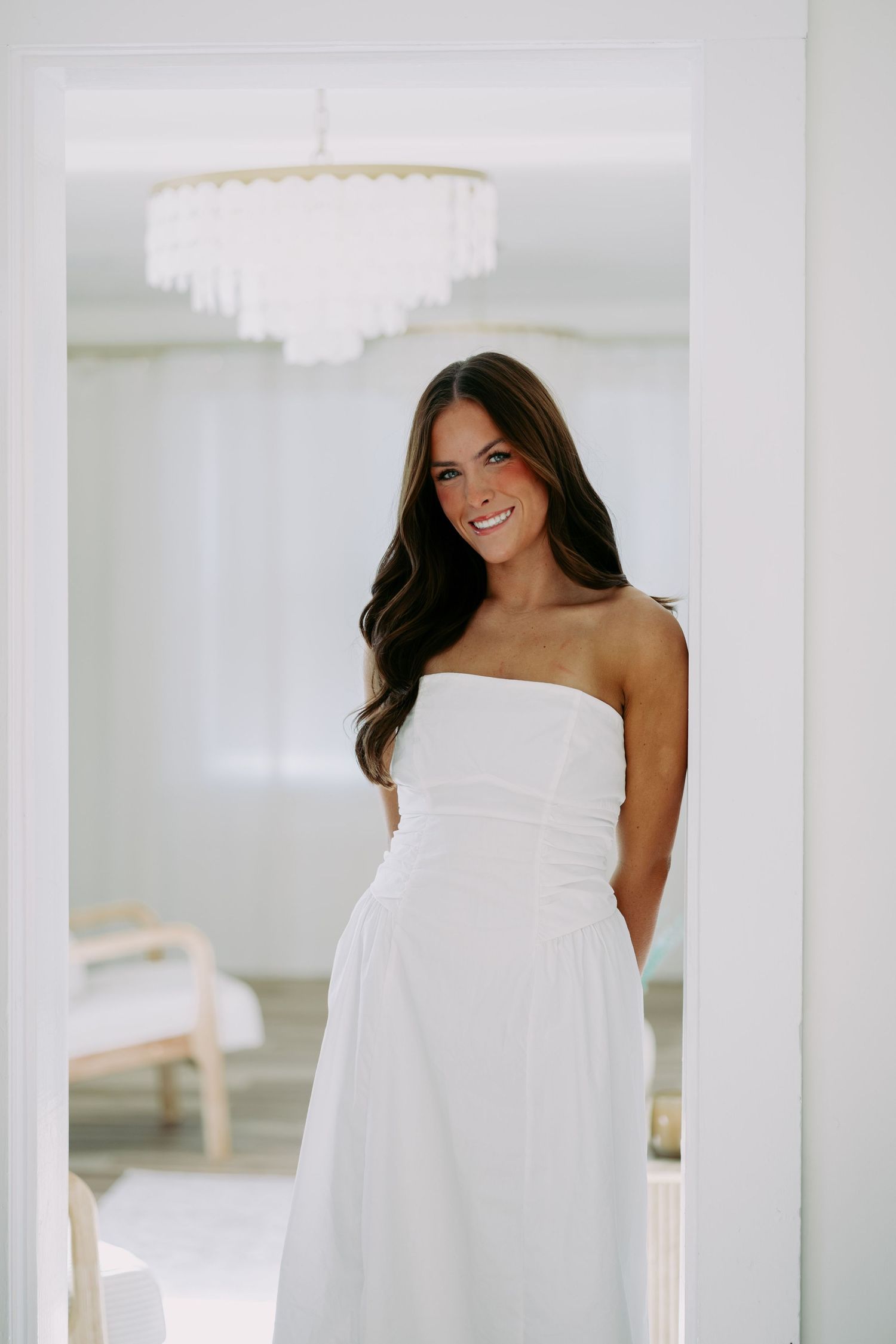 Brunette woman in white strapless maxi dress smiling in elegant white room doorway with chandelier.
