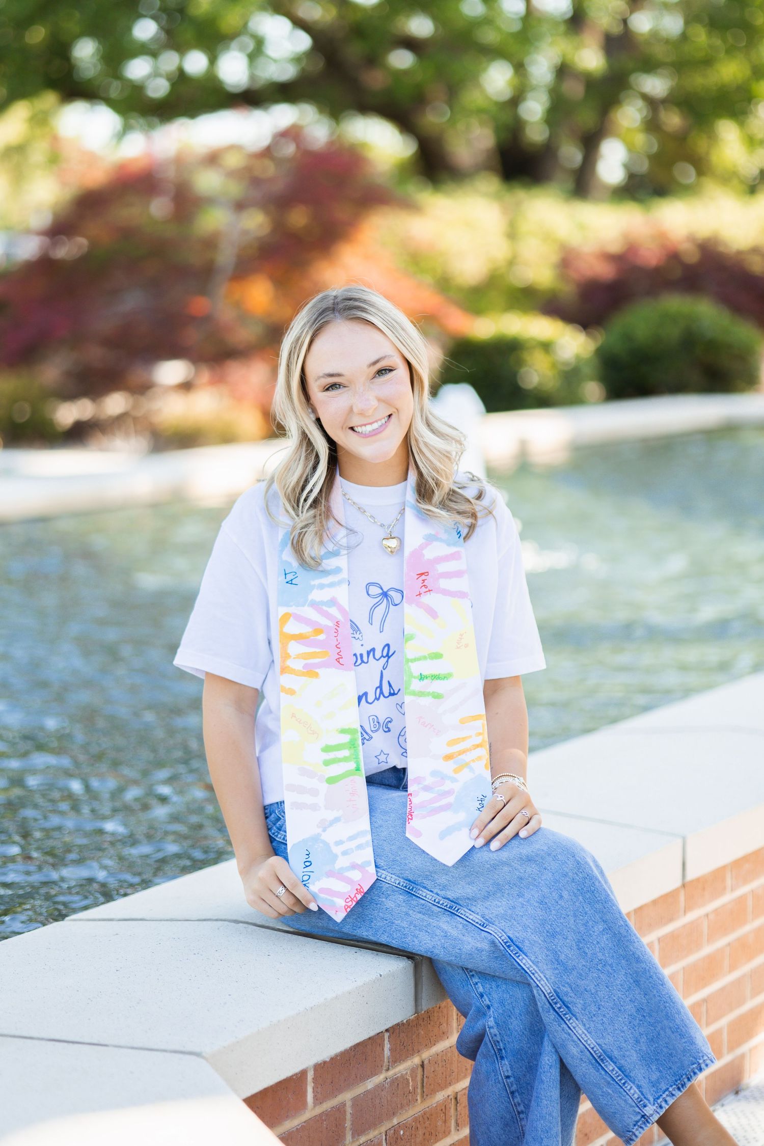 Smiling blonde graduate wearing colorful handprint graduation stole seated by campus fountain.